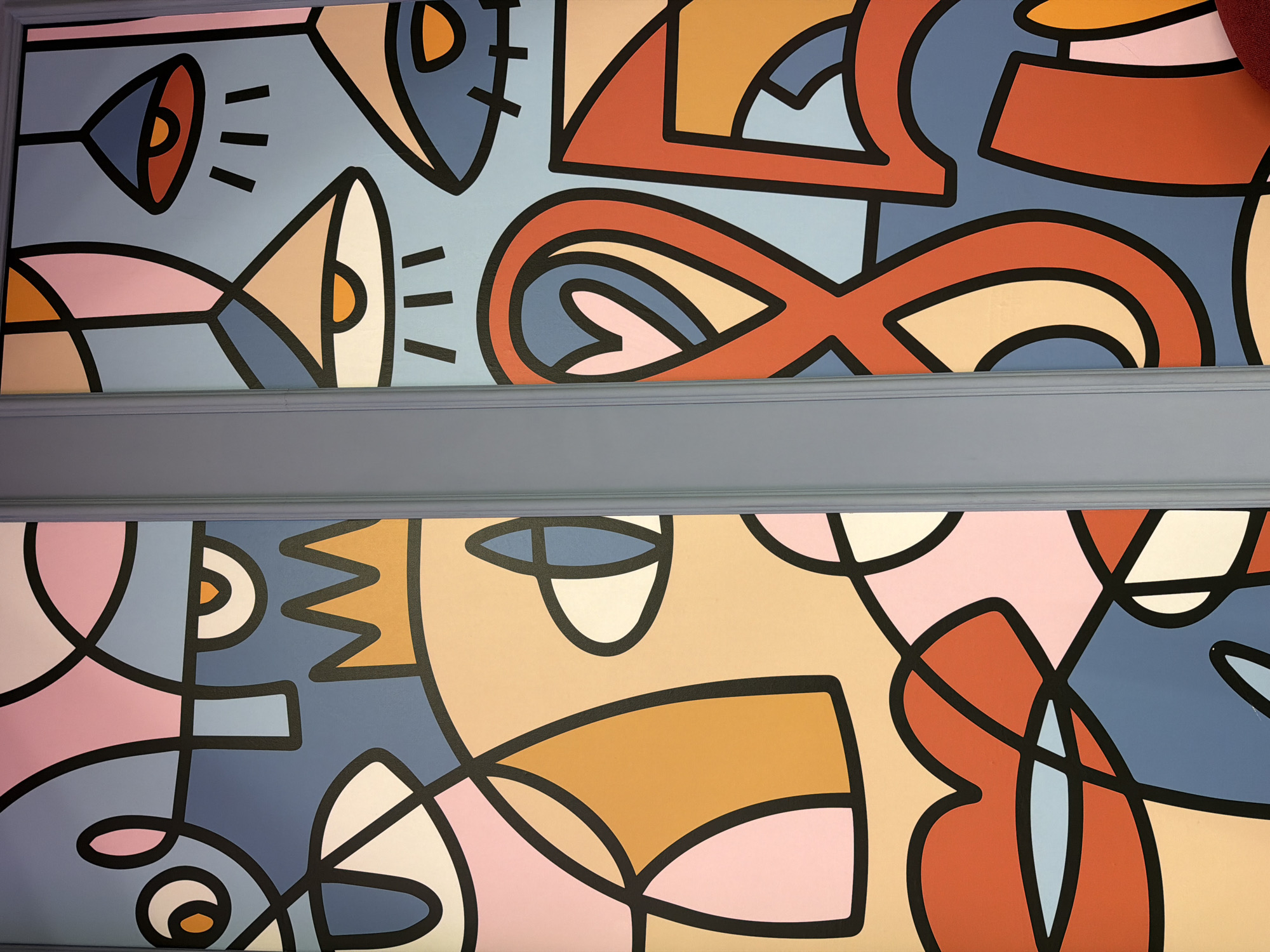 The image shows a section of a wall or ceiling with an abstract mural. The mural is divided into two sections by a gray molding. The upper section features a color palette of blues, reds, and yellows. The lower section utilizes pinks, blues, tans, and reds. The design in both sections is characterized by bold black outlines forming abstract shapes. No people are visible. There is no location in Moscow, Russia visible in the image.