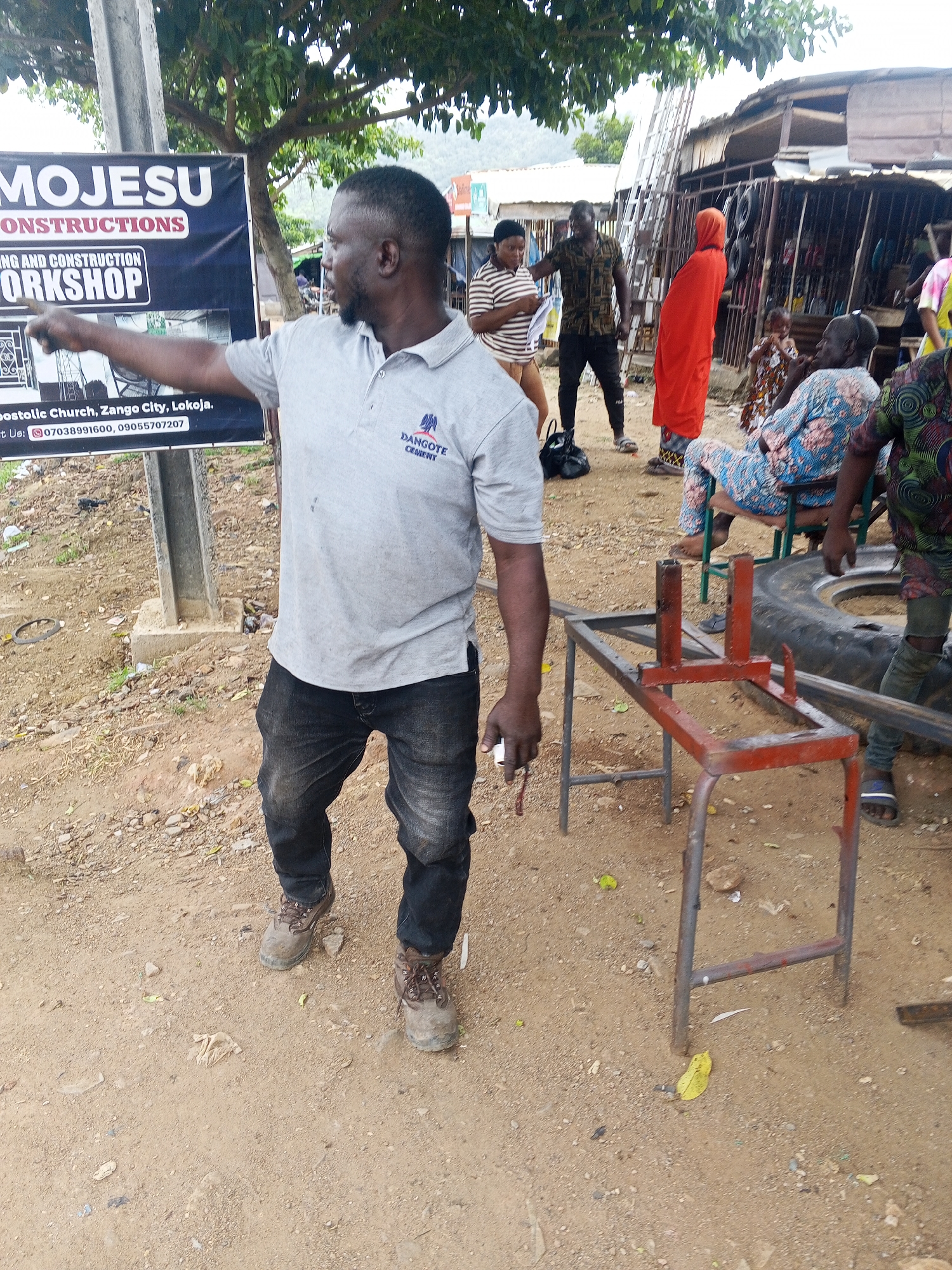 In Zango-ndaji, Nigeria, a man in a grey polo shirt and dark jeans gestures towards a banner advertising "MOJESU CONSTRUCTIONS" and a "WORKSHOP" at "Apostolic Church, Zango City, Lokoja." He is standing on a dirt ground with scattered debris. Behind him, several people are gathered near a row of structures that appear to be market stalls or workshops. One woman in a striped shirt holds documents, and a man in a patterned shirt stands beside her. Another woman in an orange headscarf and dress is visible, and a child in a patterned dress is near her. Further back, a person in a blue and pink patterned outfit is seated, and another person in a green and black patterned shirt is standing. A metal frame with red accents, possibly a piece of equipment, is in the foreground, and a large tire is visible to its right. The environment is outdoors, with trees and a cloudy sky.
