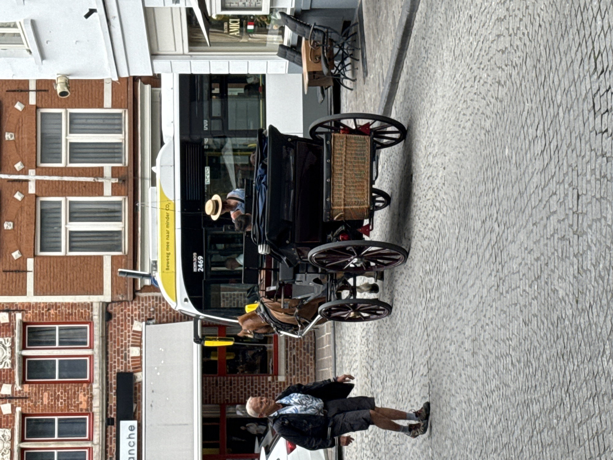 The image captures a street scene in Bruges, Belgium, likely near a historic center given the architecture. In the foreground, an elderly man with short white hair is walking from left to right, wearing a patterned short-sleeved shirt, dark shorts, and athletic shoes. He is looking towards the right. To his right, a horse-drawn carriage with a black frame and a woven basket attached to the rear is parked. The horse is a light brown color. Inside the carriage, a person wearing a straw hat and a blue top is seated. A bus with the number "2469" visible on its front is parked behind the carriage. The bus has a yellow stripe and text reading "Beweeg mee naar minder CO₂". In the background, there are brick buildings with traditional windows, and a cafe or restaurant with outdoor seating is visible on the right side of the frame, featuring chairs and tables. The street is paved with cobblestones. The overall lighting suggests it is daytime, possibly overcast.