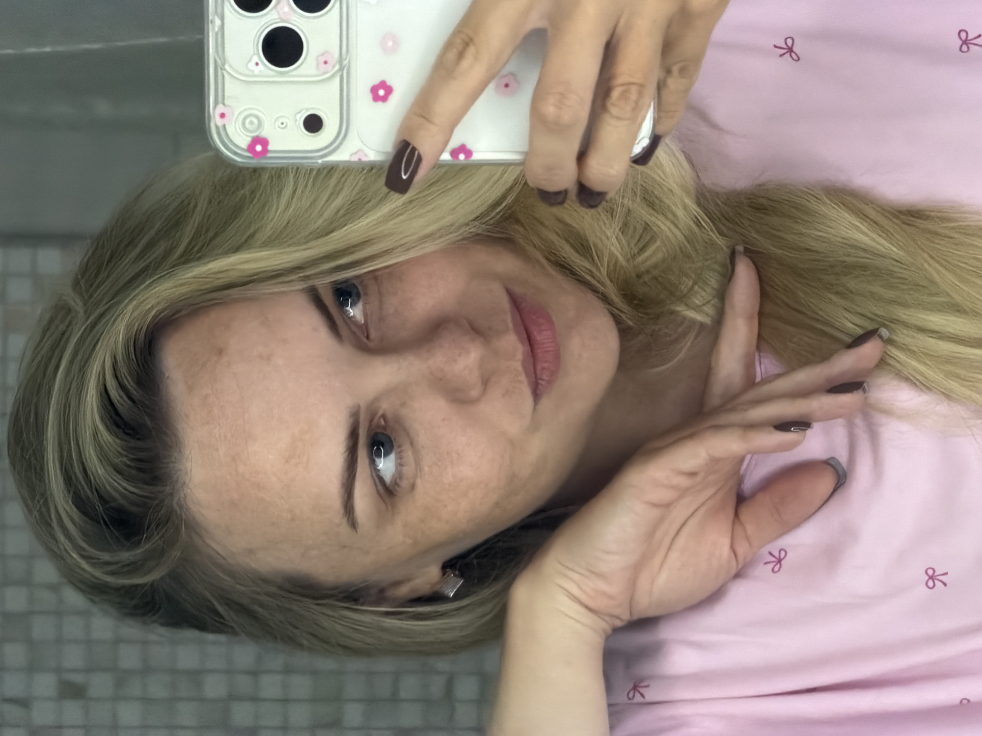 A blonde woman with freckles takes a selfie in what appears to be a bathroom. She is wearing a pink shirt with a bow pattern and has dark brown nail polish on. Her right hand is holding a white phone case with pink floral designs. Her left arm is resting on her chest, and her hand is positioned near her face. The background shows a tiled wall and a mirror. There is no indication of the city of Votkinsk, Russia.