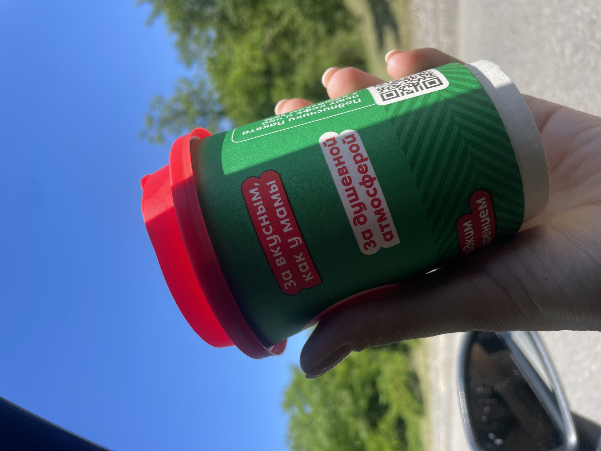 A hand holds a green disposable cup with a red lid against a bright blue sky with blurred green trees in the background. The cup has text printed on it in Russian, indicating it's for "tasty, like mom's" and "for a soulful atmosphere." The time of day appears to be daytime with clear weather. The hand holding the cup is partially visible, with manicured fingernails. The overall scene suggests a casual outdoor moment. Text on the cup is partially visible and reads "адким роением", "За вкусным, как у мамы", "За душевной атмосферой", and "Подписчики п акета и ча".