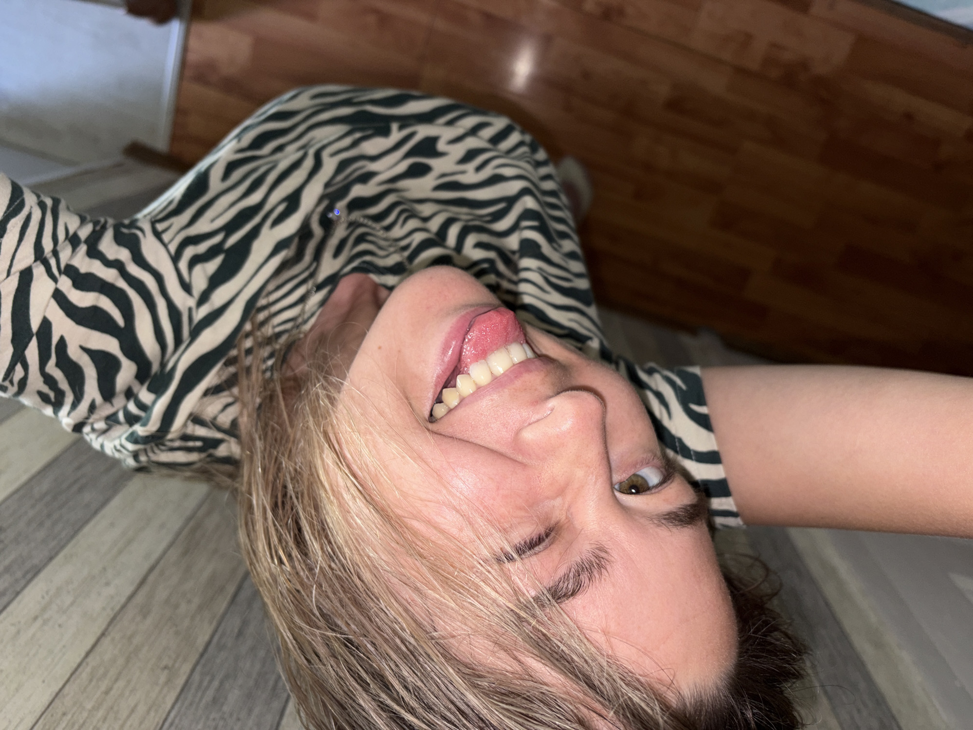 An individual is shown in a close-up, angled shot, with their head tilted. They are wearing a dress with a black and white zebra print. Their eyes are open, and their tongue is sticking out. The background consists of a wooden floor and a wall with horizontal paneling. There is no identifiable information linking the image to Chaikovsky, Russia.