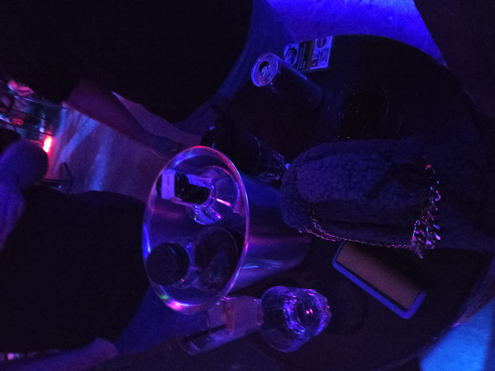 The image shows a dimly lit table with various items. There are several glasses, some of which are empty, along with a can of beverage. A small purse or bag is also present on the table. The environment appears to be a bar or club setting, given the lighting and presence of alcoholic beverages. The location within the city of Unknown, Unknown is not specified in the image.
