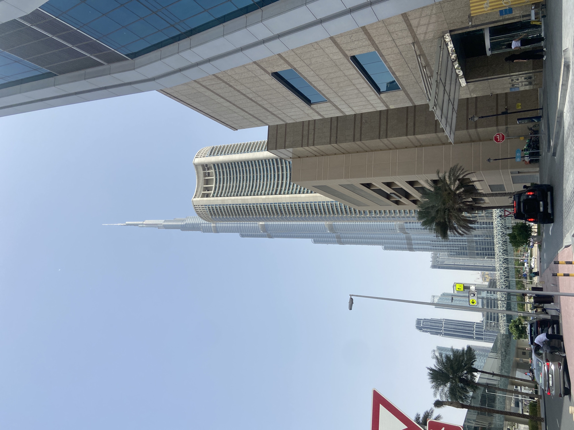 The image shows a street view in Dubai, United Arab Emirates, with the Burj Khalifa prominently visible in the background. The camera is pointed upwards, capturing a mix of modern architecture. Various buildings are visible in the frame, along with street signs and palm trees. A black car is seen on the street, and there are people near a building entrance. The sky is clear and light blue.