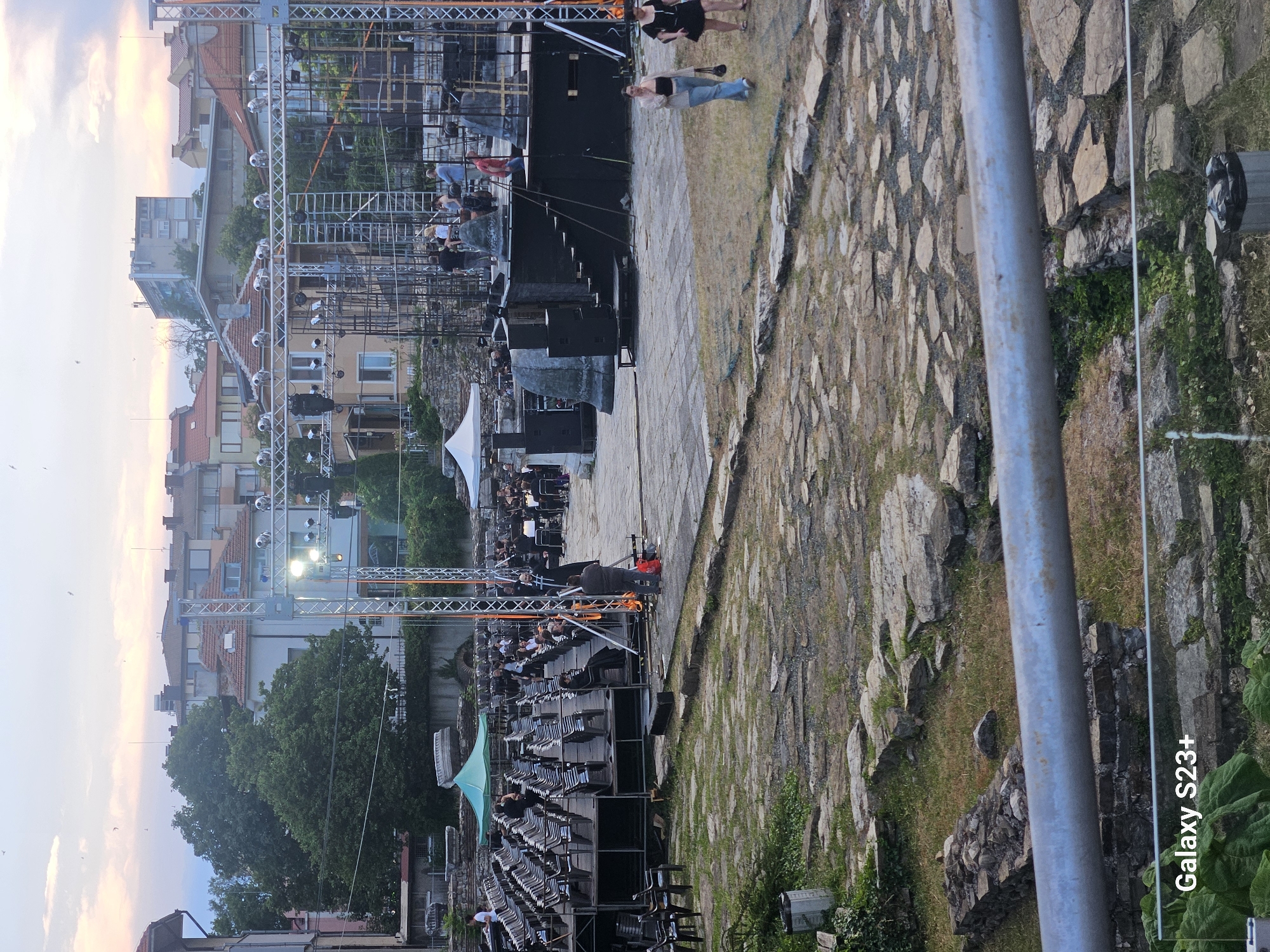 This image captures an outdoor event with a stage setup and seating area, likely a concert or performance. The scene is set in Stara Zagora, Bulgaria, with buildings visible in the background, suggesting an urban or semi-urban location.
The main subjects are a large stage structure made of metal scaffolding, adorned with lighting equipment, and rows of empty chairs arranged in front of it. Several people are present, some standing near the stage and others seated in the audience area, though the crowd is not dense. The overall atmosphere appears relaxed, with a casual gathering for an event.
The time of day is likely late afternoon or early evening, as indicated by the soft, diffused light and the warm hues in the sky. The weather appears to be fair, contributing to the outdoor event. The ground is a mix of paved stone and grass, with some greenery around the edges, hinting at a park or historical site.
Notable details include a white tent structure to the left of the stage and various sound and lighting equipment scattered around. A prominent metal pole runs along the right side of the image. The "Galaxy S23+" text is visible at the bottom right, indicating the device used to capture the photo. The event seems to be in its initial stages or winding down, given the partially filled seating.