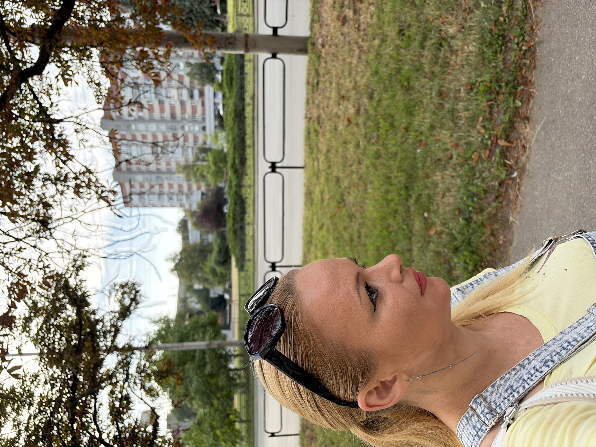 A medium shot captures a blonde woman in profile, looking to the right. She is wearing a yellow t-shirt and denim overalls, with black sunglasses perched on her head. Her hair is pulled back into a ponytail, and she has noticeable winged eyeliner and pink lipstick. A long, delicate earring dangles from her left ear.
The background suggests an urban environment. A tree with autumn leaves partially obscures the view, revealing a multi-story apartment building in the distance. A metal fence separates a grassy area from a road. The lighting indicates it is daytime, possibly late afternoon.