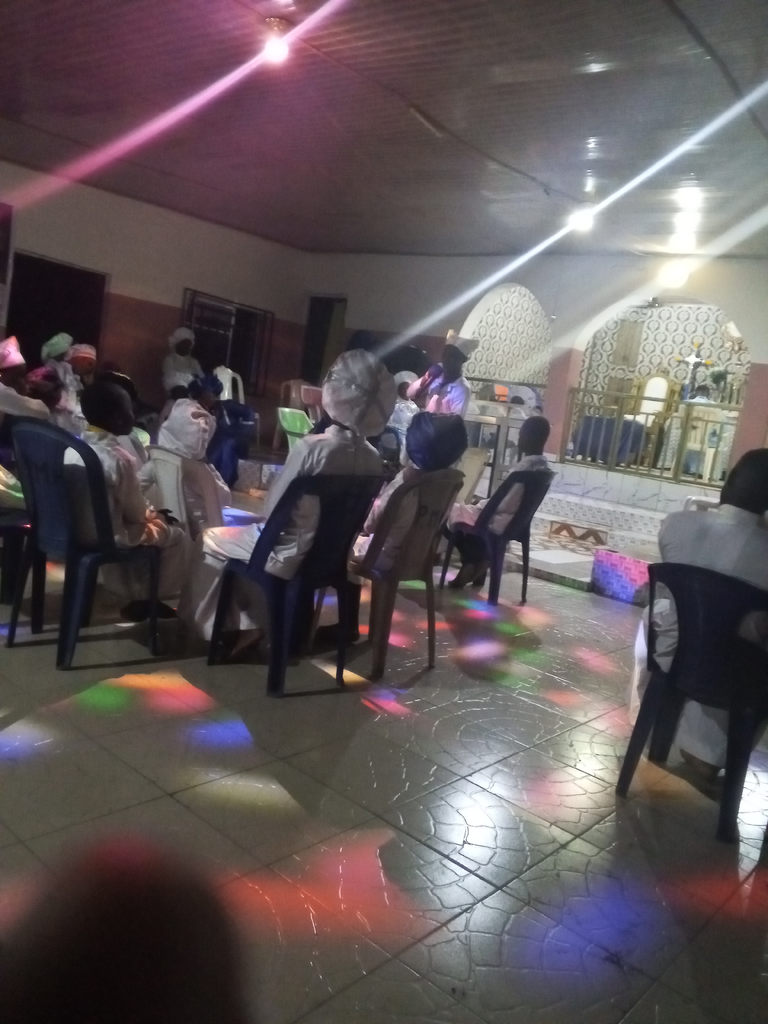 A gathering of people, mostly dressed in white attire, is taking place indoors in Lokoja, Nigeria. The individuals are seated on chairs, facing an elevated area that appears to be a stage or platform decorated with white and blue patterns. The floor has a tiled surface, illuminated with colorful light patterns. The room is lit by artificial lighting.