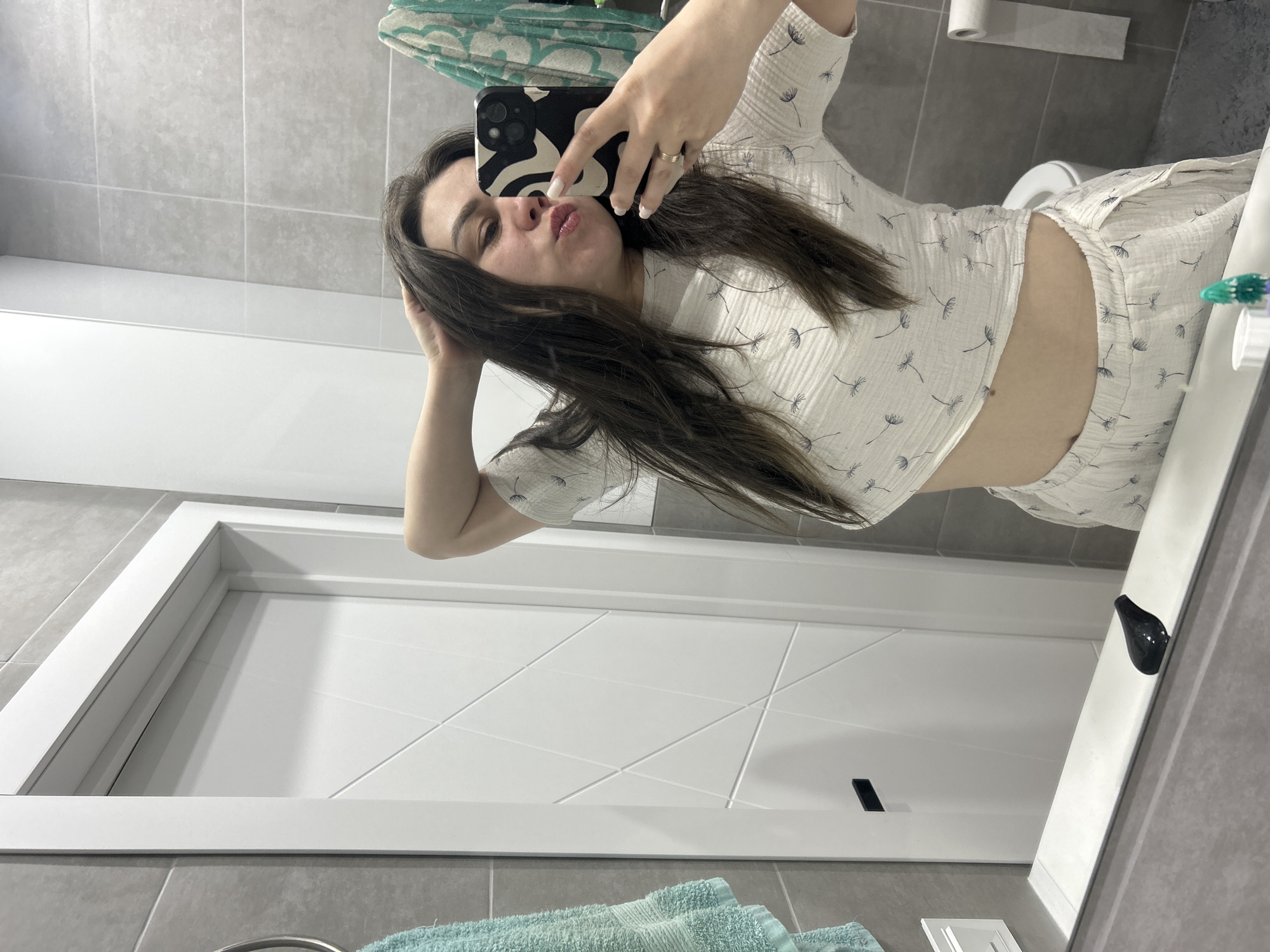 A woman in a white patterned short set takes a selfie in a bathroom. She is holding a black and white patterned phone. She has long brown hair. Behind her are gray bathroom tiles, a white door, a toilet, and a green towel. The location is Chaikovsky, Russia.