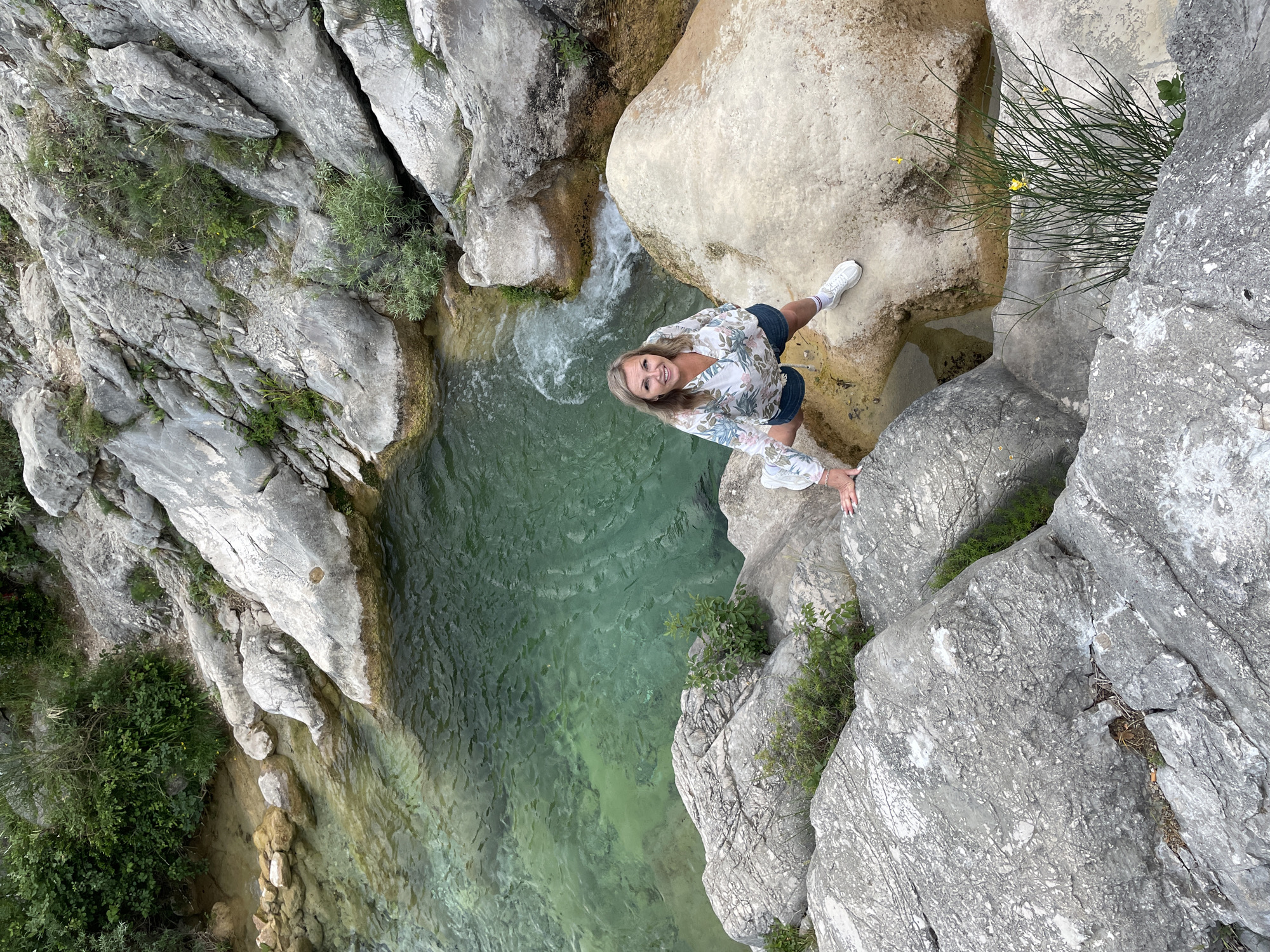 The image depicts a breathtaking natural landscape in Zupci, Montenegro, showcasing a deep, rocky gorge or canyon with a clear, emerald-green natural pool or stream flowing through it. The perspective is an aerial shot, looking directly down into the canyon, emphasizing its depth and the pristine quality of the water.
The dominant features are the rugged, light grey rock formations that form the steep sides of the gorge. These rocks exhibit varied textures and crevices, where resilient green vegetation, including shrubs and grasses, thrives. In the upper middle section, a small, frothy cascade indicates the flow of water into the main pool.
A woman with blonde hair is positioned on a rocky ledge in the mid-right of the frame. She is smiling broadly and looking up towards the camera. She is dressed in a light-colored, long-sleeved floral blouse, denim shorts, and white sneakers. Her right hand rests on the rock in front of her, suggesting a pose or a moment of balance while enjoying the view.
The scene takes place during bright daylight, indicative of clear and pleasant weather, ideal for outdoor activities. The woman's cheerful expression conveys joy and appreciation for the stunning natural environment. The vibrant green of the water suggests a clean, possibly freshwater, source. There is no visible text in the image.