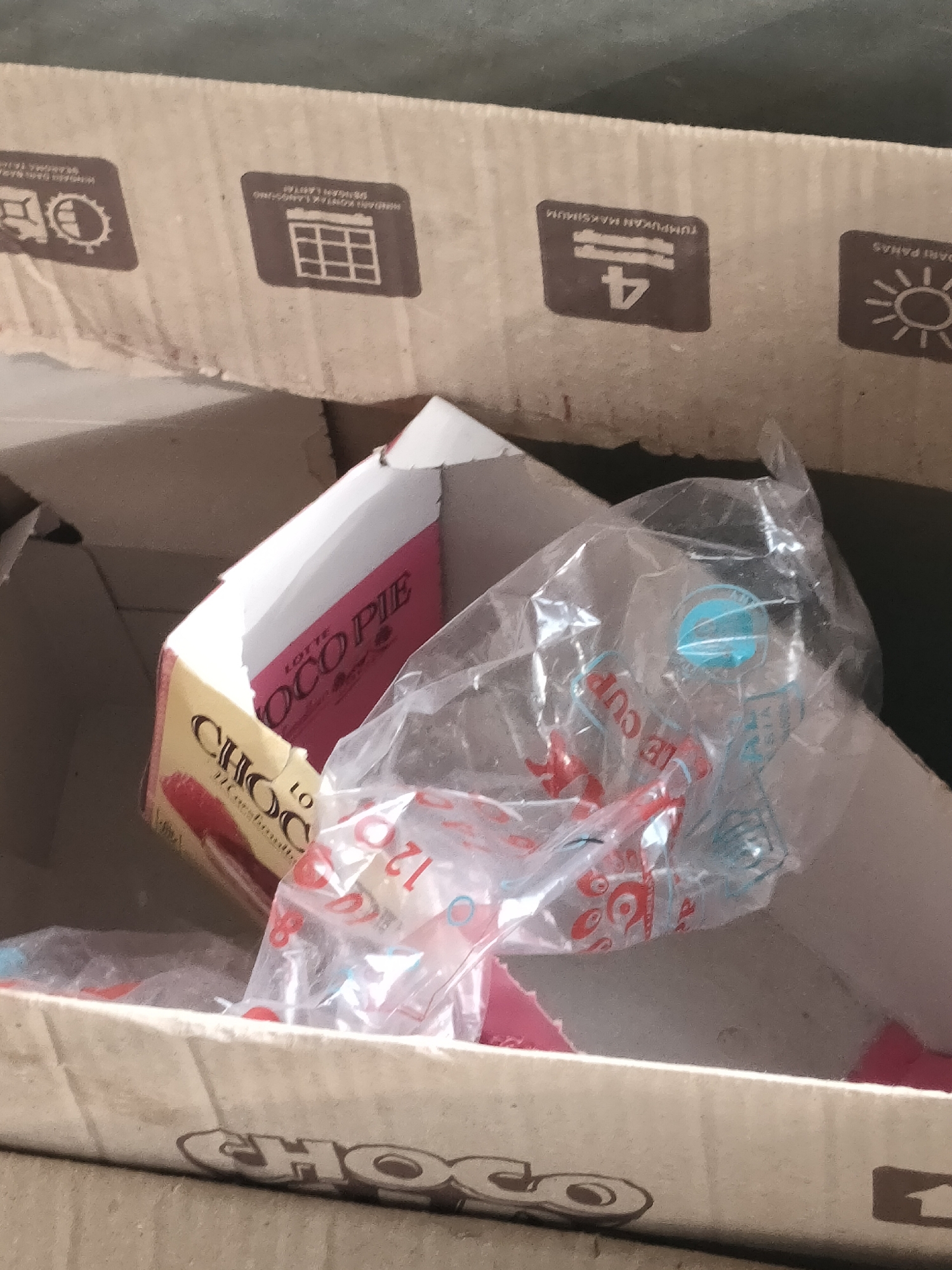 The image shows a crumpled cardboard box containing a few individual servings of Lotte Choco Pie. The box is slightly open, revealing the pink and yellow packaging of the dessert. Inside the box, there is also some clear plastic packaging, possibly from additional items or wrapping. Some of the cardboard exterior of the box has printed icons, including a calendar, a number 4, and a sun with rays. The scene appears to be indoors, with a muted, slightly textured background. The lighting is indirect. The image context suggests the box might have been recently opened or is in a state of disarray.