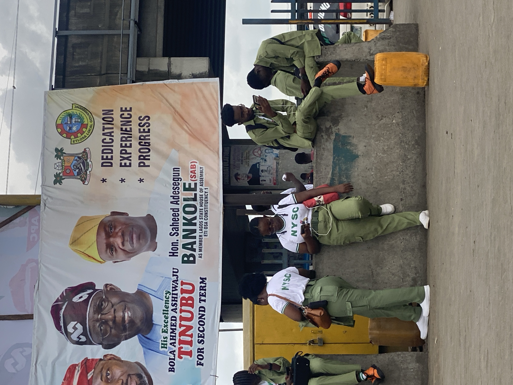 This image captures an outdoor scene in an urban area of Lagos, Nigeria, likely during the daytime under an overcast sky. Several young adults, identifiable as National Youth Service Corps (NYSC) members by their distinctive green trousers, white shirts with "NYSC" branding, and sometimes matching caps, are present.
The NYSC members are primarily seen resting or waiting. Two male corps members are seated on a concrete ledge or steps, both looking at their mobile phones. One of them is wearing orange and black sneakers, while the other has black shoes. Further along the ledge, a female corps member is lying down, also engaged with her phone, next to a yellow jerrycan. Another female corps member stands nearby, carrying a brown purse. One male corps member at the top right of the frame is seated on the steps, while another at the bottom is partially visible. One of the yellow jerrycans is being used as a makeshift seat.
A large political banner dominates the left side of the frame, featuring portraits of politicians. Prominently displayed are "His Excellency BOLA AHMED ASHIWAJU TINUBU FOR SECOND TERM" and "Hon. Saheed Adesegun BANKOLE (SAB) AS MEMBER LAGOS STATE HOUSE OF ASSEMBLY ETI OSA CONSTITUENCY 1". The banner also shows the emblem of "ETI-OSA LOCAL GOVERNMENT" and the slogan "DEDICATION EXPERIENCE PROGRESS". Other partial texts like "AKA SEGO" and "BURTH" are visible. The background features a concrete wall, some dark metal structures, and part of a building.