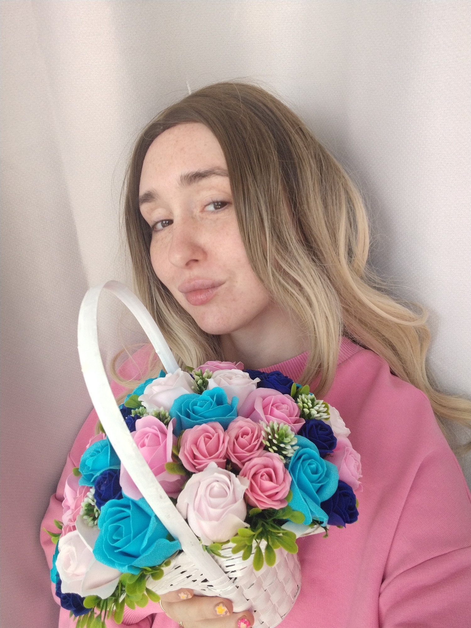 A female individual is depicted from the waist up, centered slightly to the right, looking directly at the camera with a slight pout. She has long, wavy light brown/blonde hair and visible freckles across her nose and cheeks. She is wearing a solid pink long-sleeved garment, possibly a sweatshirt or sweater. She holds a white wicker basket containing an arrangement of artificial flowers, predominantly roses, in shades of light pink, dark pink, turquoise, dark blue, and white, intermingled with small green foliage elements. Her right hand, adorned with a ring on one finger and a small yellow star decal on a fingernail, is visible gripping the basket. The background consists of a plain, light-colored interior wall, appearing to be a corner where two walls of subtly different light tones meet. The specific location within Izhevsk, Russia, is not discernible from the visual evidence.