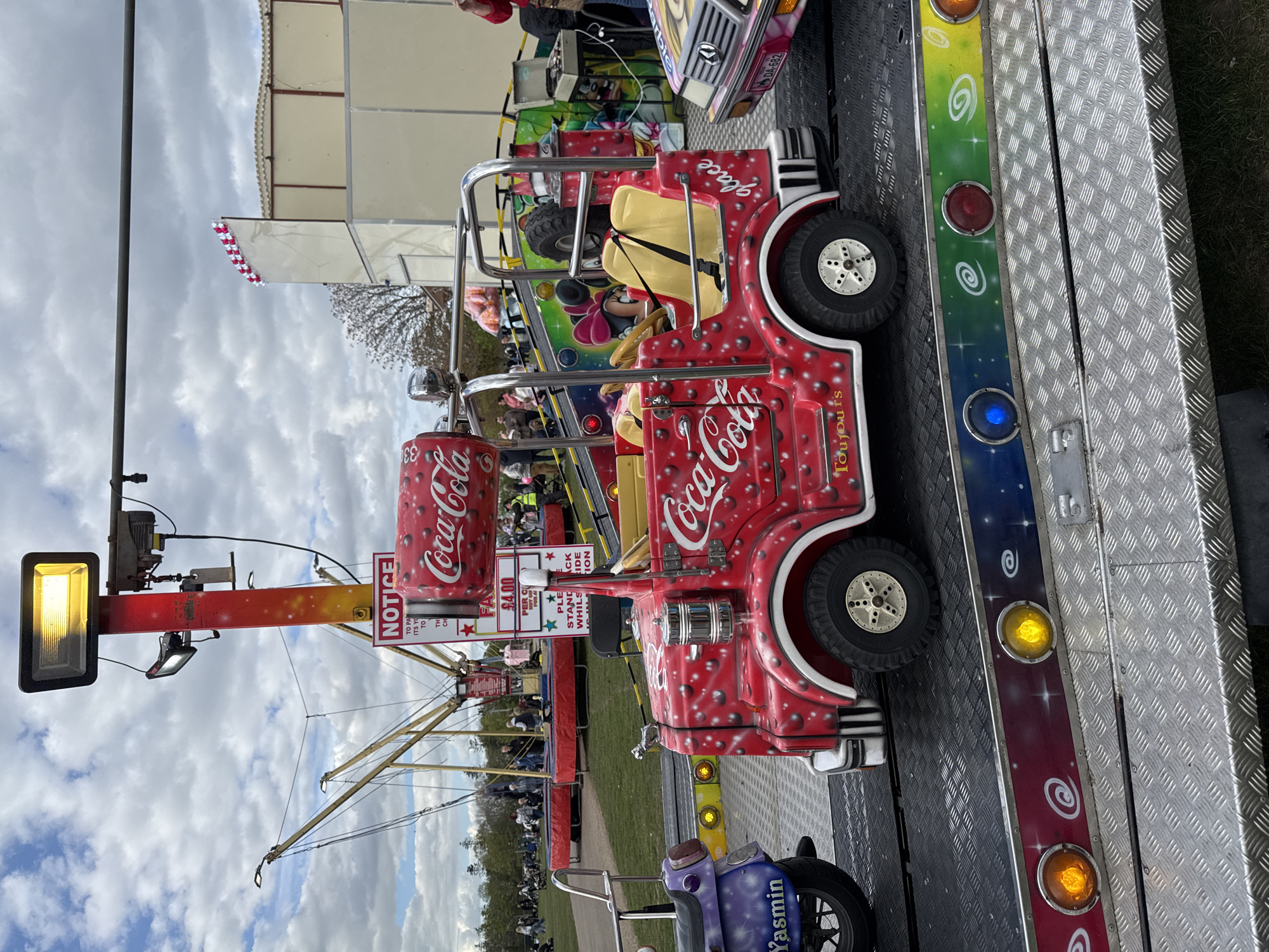 The image depicts a children's fairground ride, specifically a circular track with vehicles and motorcycles. A prominent feature is a red car adorned with Coca-Cola branding. Other vehicles, including a purple motorcycle labeled "Yasmin," are visible. There's a large crowd of people in the background, suggesting a busy fairground atmosphere. A notice sign is adjacent to the red Coca-Cola car and some metal support arms extend over a grassy area, and lighting fixtures are attached to a pole. The scene is likely in South Norfolk, United Kingdom based on the image analysis.