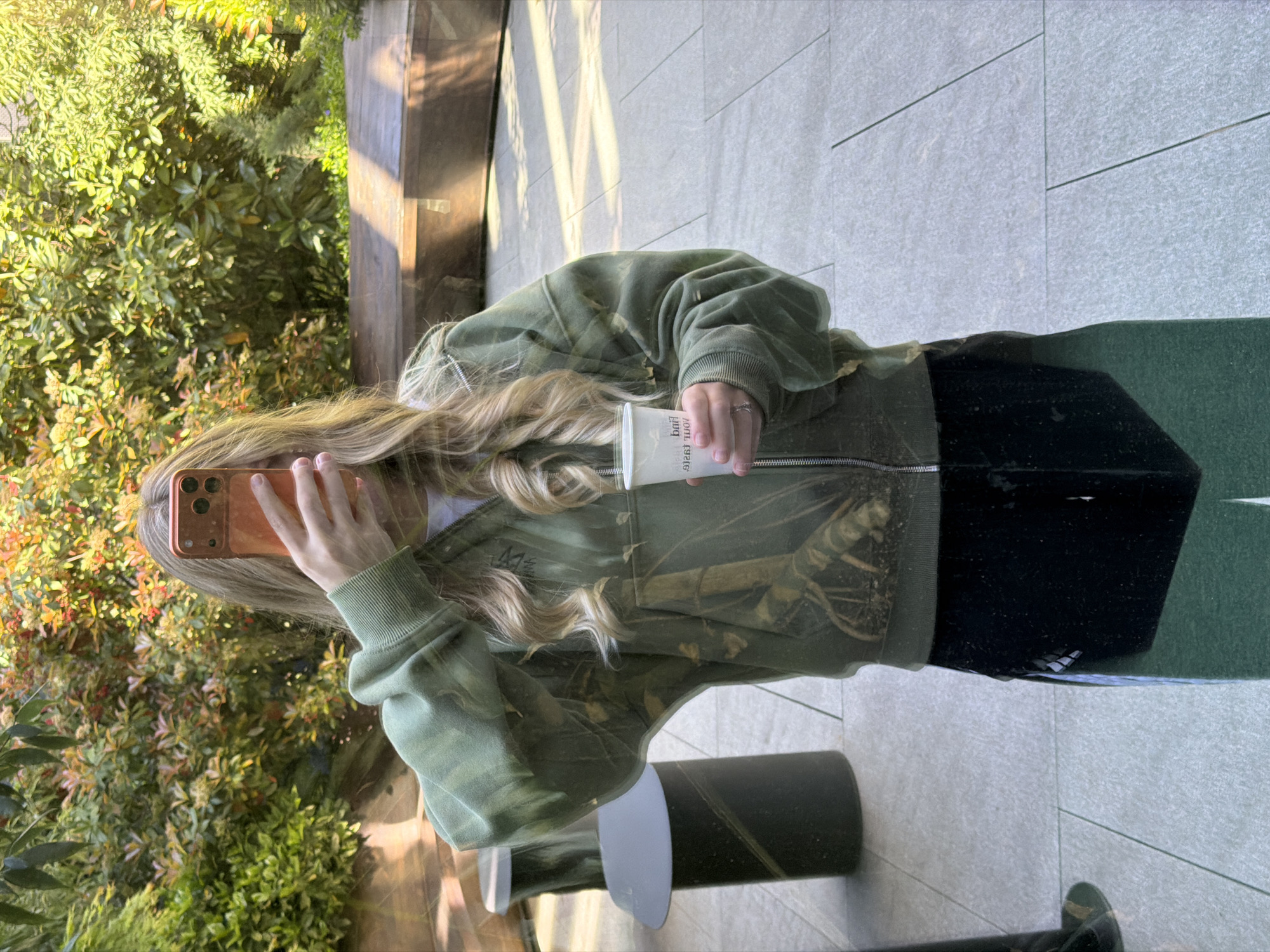 The image displays a person standing outdoors on a bright, sunny day. The individual has long, wavy blonde hair and is wearing a green zip-up hoodie with a subtle patterned design, a white t-shirt underneath, and dark green or black pants/skirt.
The person holds an orange-gold smartphone in their left hand, with the screen facing them, obscuring their face. In their right hand, they hold a white cup. Text is visible on the cup, appearing to be "Pink Cola," although it is partially obscured or mirrored.
The background on the left is filled with lush green foliage, including various bushes and trees, suggesting a natural or landscaped area. To the right, there is a light-colored tiled or paved surface, which could be a floor or a wall, and a dark, cylindrical object or pillar. Strong sunlight casts prominent shadows across the scene, indicating clear weather. The setting appears to be a casual outdoor location, possibly in Tbilisi, Georgia, as indicated by the context. The image captures a candid, everyday moment.
