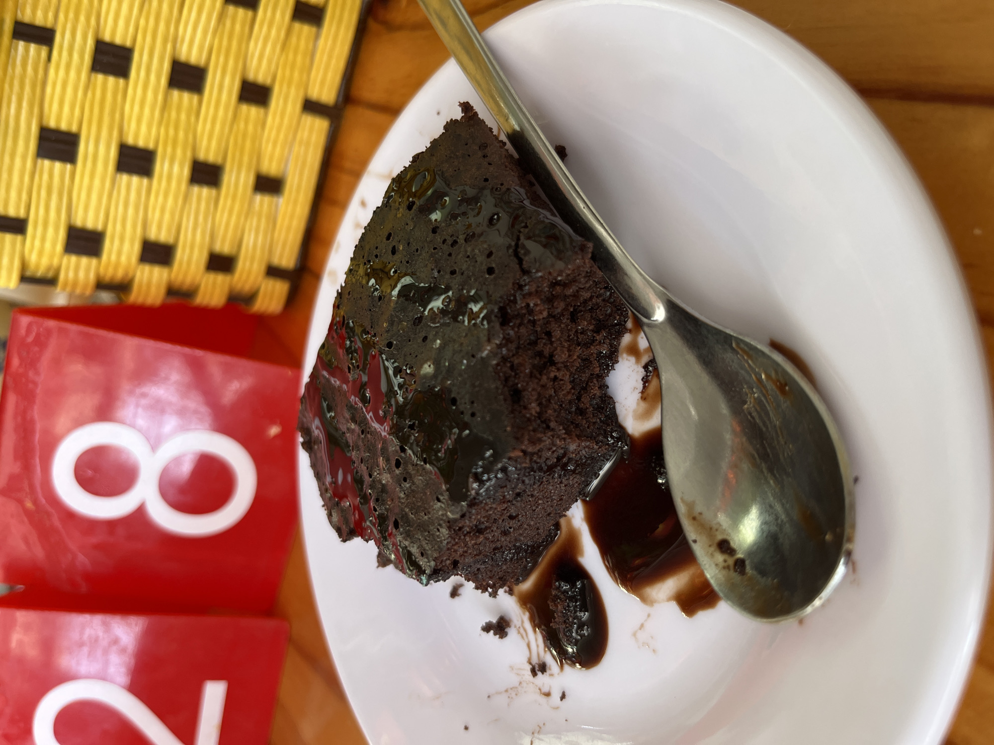 A close-up shot captures a piece of chocolate cake on a white plate, accompanied by a spoon. Dark chocolate sauce is visible on the cake and plate. A red object with the number "8" is partially visible on the left side of the frame, and a yellow woven object is in the background. The scene appears to be set indoors on a wooden surface.