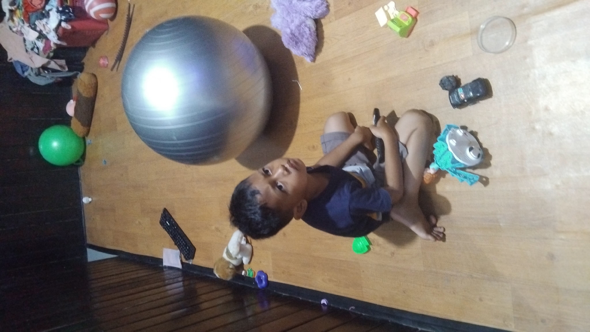 In Bangun Rejo, Indonesia, a young boy of approximately 5-7 years old is seated on a wooden floor in what appears to be a room. He is wearing a dark blue t-shirt and grey shorts. The boy is holding a black remote control in his right hand and looking upwards and to his right. Scattered around him are various objects, including a large, silver exercise ball, a green bouncy ball, a small plush toy resembling a dog, a purple plush toy, a toy vehicle, and a small toy with a turquoise cloth. In the background, a laundry basket filled with clothes is visible, along with a portion of a wooden wall. The lighting in the room is dim, suggesting it may be evening or the room is not well-lit.
