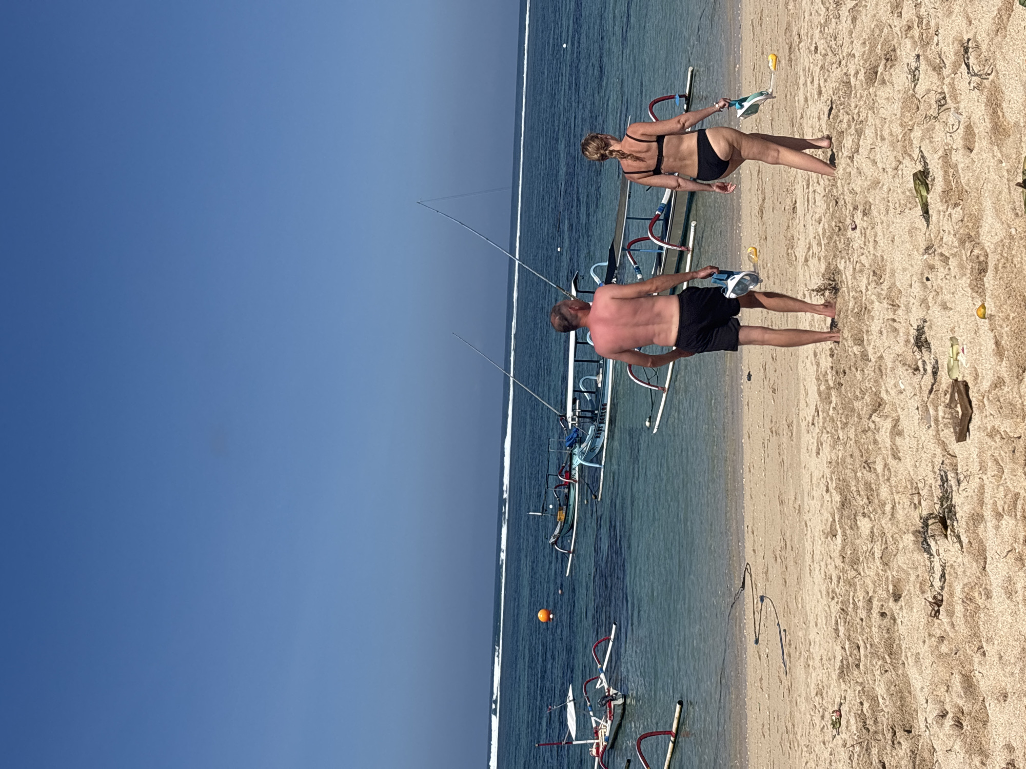 In Kuta Selatan, Indonesia, a man and a woman are walking on a sandy beach. The man, with tanned skin and short dark hair, wears black swim shorts. The woman, with blonde hair braided down her back, wears a black bikini. Both are holding snorkeling masks. In the background, several traditional outrigger boats are moored in clear turquoise water. The ocean has a thin white line of breaking waves in the distance, under a bright blue sky. The beach is covered in light brown sand, with scattered dry seaweed and debris.