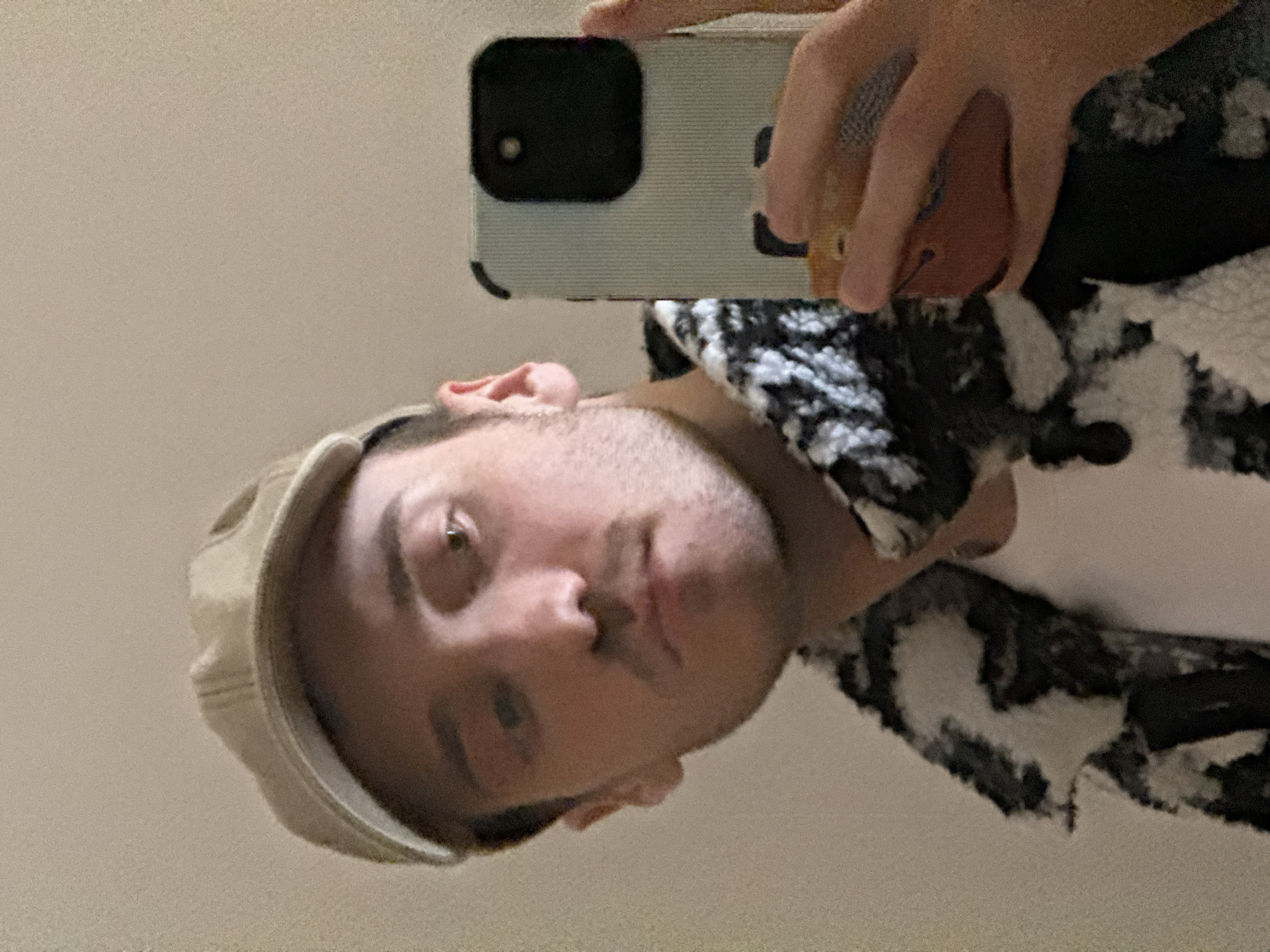 A male individual is captured in a close-up selfie, taken either with a smartphone's front camera or reflected in a mirror. He is centered in the frame, wearing a beige cap, a white t-shirt, and a black and white patterned, possibly fleece or sherpa, jacket. His right hand holds a smartphone, partially obscuring his lower right jaw and shoulder. The smartphone features a light blue, vertically ribbed case with a distinct black square camera module. The individual has short dark hair, light brown eyes, and stubble on his face. His expression is neutral. The background consists of a plain, light-colored, likely interior wall. The exact location in the city of Kazan, Russia, cannot be determined from the visual information provided within the image.