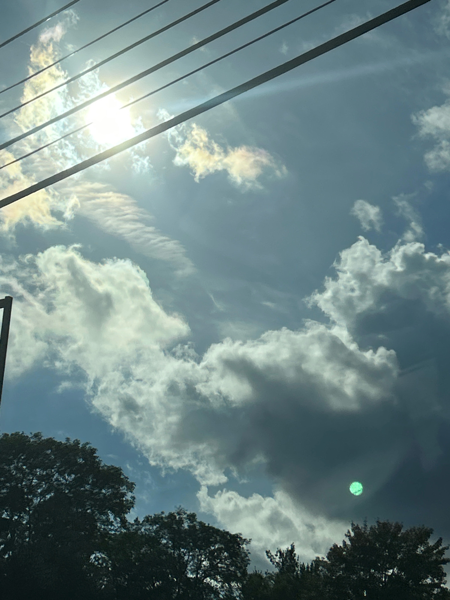 This is a photograph taken in Collier Township, United States of America, featuring a sky filled with clouds and sunlight filtering through power lines. The image shows a dark silhouette of trees along the bottom edge, suggesting it was taken from ground level looking upwards. In the sky, bright sunlight beams through a gap in the clouds, creating a lens flare effect and illuminating some of the cloud formations with a yellow-white hue. Other clouds appear darker and more voluminous. A notable feature is a circular, glowing green orb in the sky, possibly an artifact of the camera or a distinct atmospheric phenomenon. The overall scene depicts a daytime sky with natural light, clouds, and man-made elements like power lines and trees.