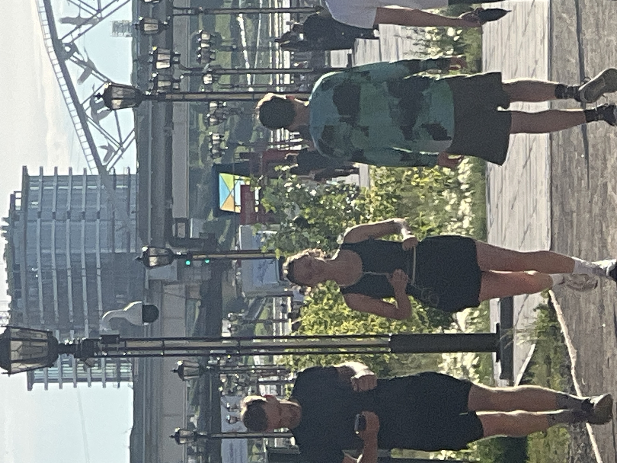 The image depicts a group of people walking and running in an outdoor urban setting. In the foreground, three individuals are visible. On the left, a man with short brown hair is jogging, wearing a black t-shirt and dark shorts, and holding a smartphone in his right hand. In the center, a woman with blonde hair is running, wearing a dark tank top and a dark skirt, and holding her arms in a running motion. On the right, a person with short dark hair is walking away from the camera, wearing a long-sleeved green shirt with a camouflage pattern and dark shorts. Further in the background, several other figures can be seen walking. The environment includes a paved pathway, lush green bushes, and multiple ornate streetlights with multiple lamps. In the middle ground, a modern glass-fronted building with multiple stories stands under a bright sky, and a large metal structure that appears to be a bridge with some decorative bird-like elements is visible. The overall scene suggests a public park or promenade. The city of Tyumen, Russia, is identified as the location of this scene.
