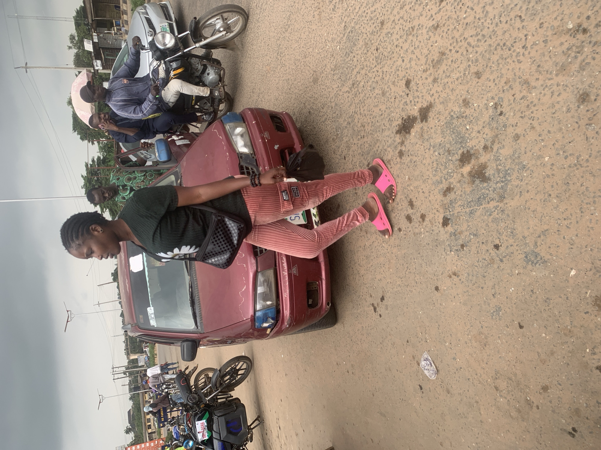 In Shagamu, Nigeria, an outdoor scene depicts a roadside with a reddish-maroon car parked on a dirt and asphalt surface. A woman, facing right, walks past the car. She has braided hair and wears a dark green shirt with a white flower design, pink striped pants, and pink slides. A black crossbody bag is slung over her shoulder, and she carries a black plastic bag in her right hand. Behind her, on the road, a motorcycle rider wearing a blue and white jacket and a black cap is visible, along with another person on the motorcycle. The background includes other vehicles, motorcycles, and structures, indicating an urban environment. The sky is overcast with a greyish hue.