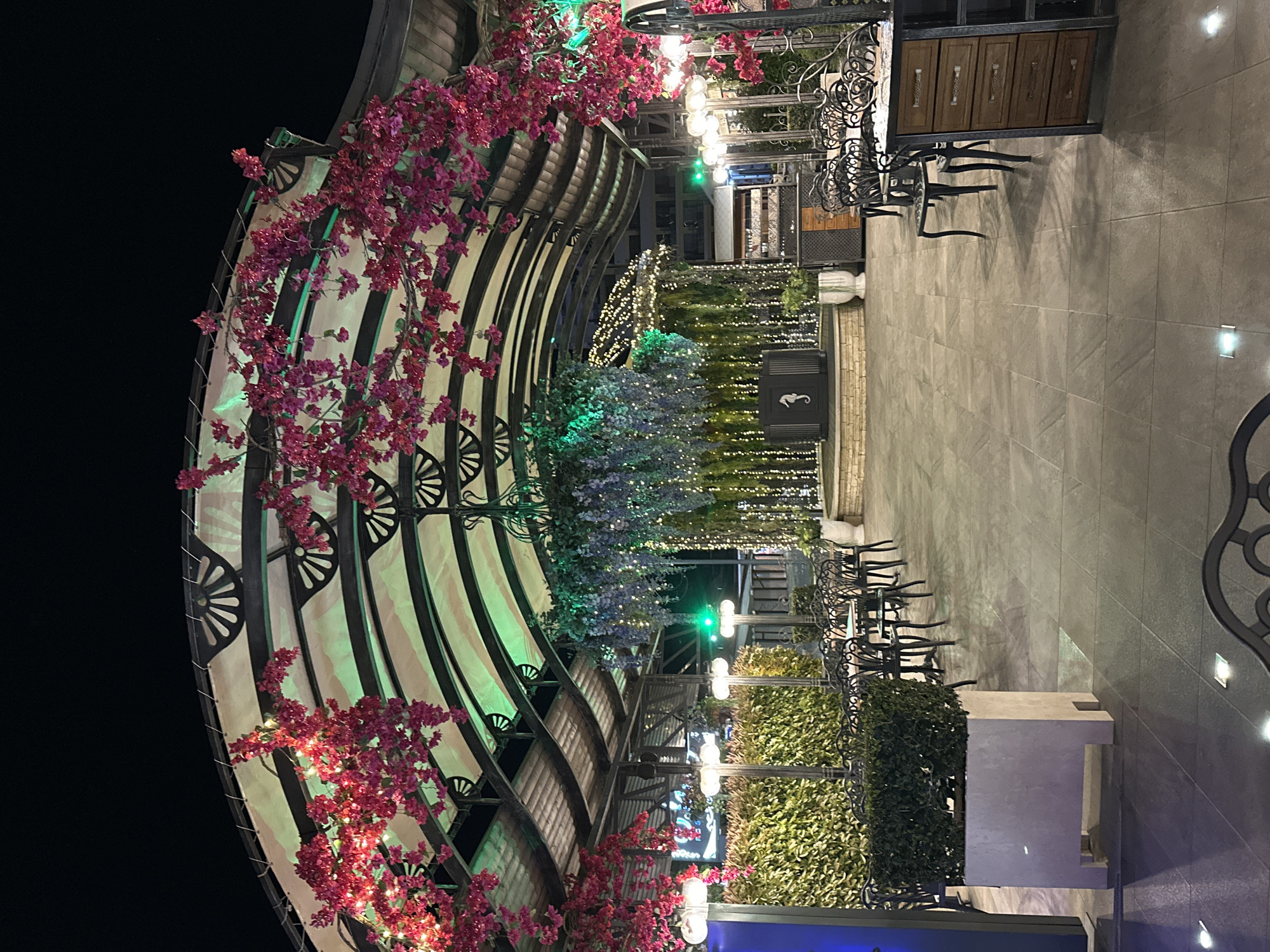 The image shows an outdoor restaurant area at night, seemingly empty of people. The scene is characterized by a curved structure made of metal ribs, adorned with pink flowers and string lights. The space is organized with tables and chairs, and the ground is paved with gray tiles. Planters filled with lush greenery and foliage are interspersed, some illuminated by lights. The lighting is a mix of string lights, green spotlights, and recessed ground lights. The sky is dark and unobscured.