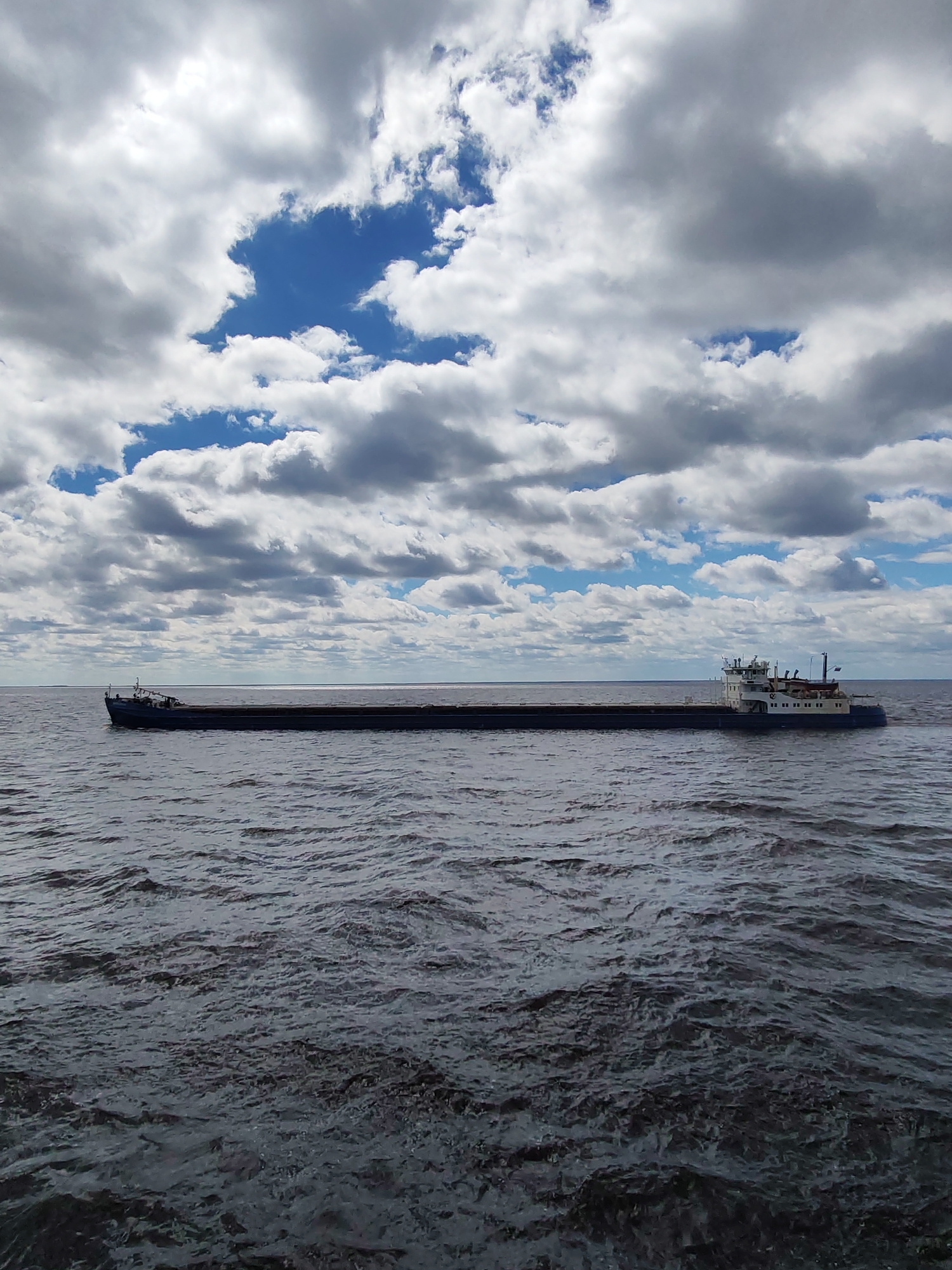 The image depicts a long, flat-bottomed barge floating on a body of water. The water is choppy with visible ripples and a dark gray hue. The barge is dark, likely a drab color, and extends nearly the width of the image, with a visible superstructure and details at each end. The sky is overcast with a mix of white and gray clouds and patches of blue. No people are visible on the boat or on the shore. It is unlikely that the location is the city of Mologa in Russia because that city does not currently exist; it was flooded in the 1940s to create the Rybinsk Reservoir. The image likely depicts a similar body of water as the Rybinsk Reservoir.