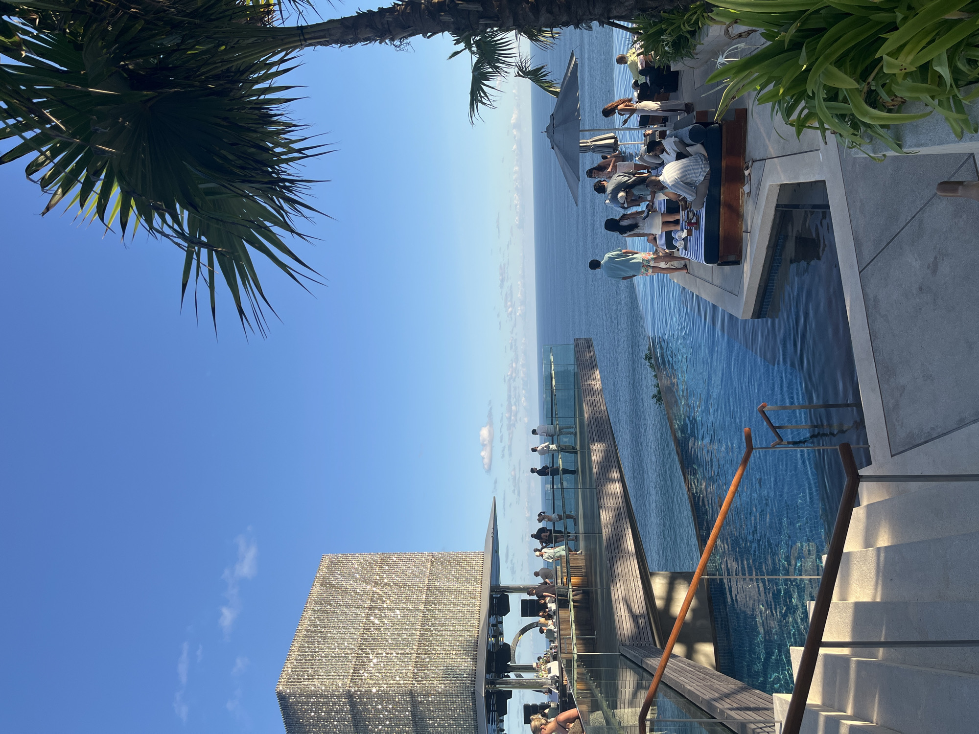 A rooftop infinity pool bar at a resort in Kuta Selatan, Indonesia, overlooks the ocean under a clear blue sky. A multi-story building with a shimmering, cubed facade is visible to the left, with people gathered on its balcony and bar area. The infinity pool extends to the horizon, with lounge chairs and umbrellas set up on a tiled deck where a group of people are relaxing. A tall palm tree frames the right side of the image, its fronds extending into the sky. The scene captures a vibrant atmosphere with people enjoying the sunset, drinks, and the stunning ocean views.