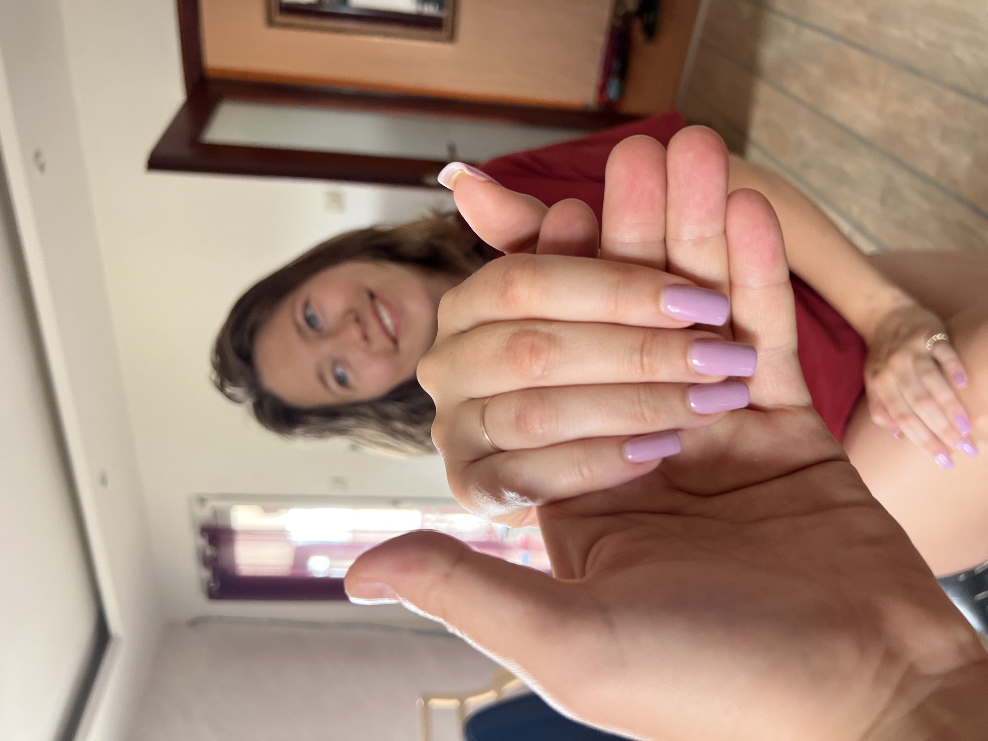 The image shows a person holding another person's hand. The person in focus has lavender nail polish on their fingernails, and is wearing a ring on one finger. The second person is blurred in the background, but appears to be smiling. The background suggests an interior setting. A doorway, wall and ceiling can be seen. The picture could be located in Podgorica, Montenegro.