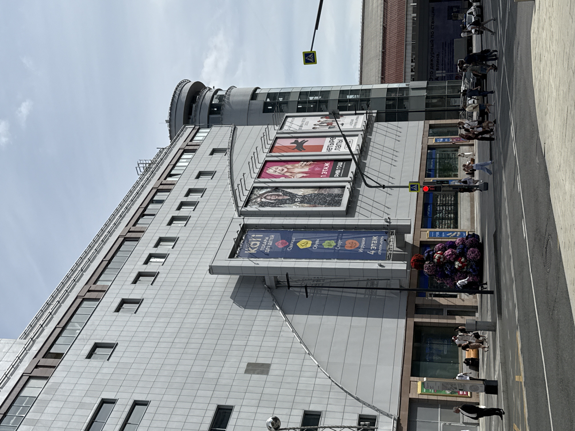 The image captures a street view in Moscow, Russia, featuring a large, modern building with a light-colored facade. The building has numerous windows and features several large billboards and banners advertising various products and brands. To the right, a cylindrical tower-like structure is attached to the main building. In the foreground, a street with pedestrian crossings and traffic lights is visible. Several pedestrians are crossing the street, and some are walking on the sidewalk. Cars are also present on the road. A prominent floral decoration, resembling a large, multi-colored cone, is situated on the sidewalk in front of the building. The sky is partly cloudy, indicating daytime.