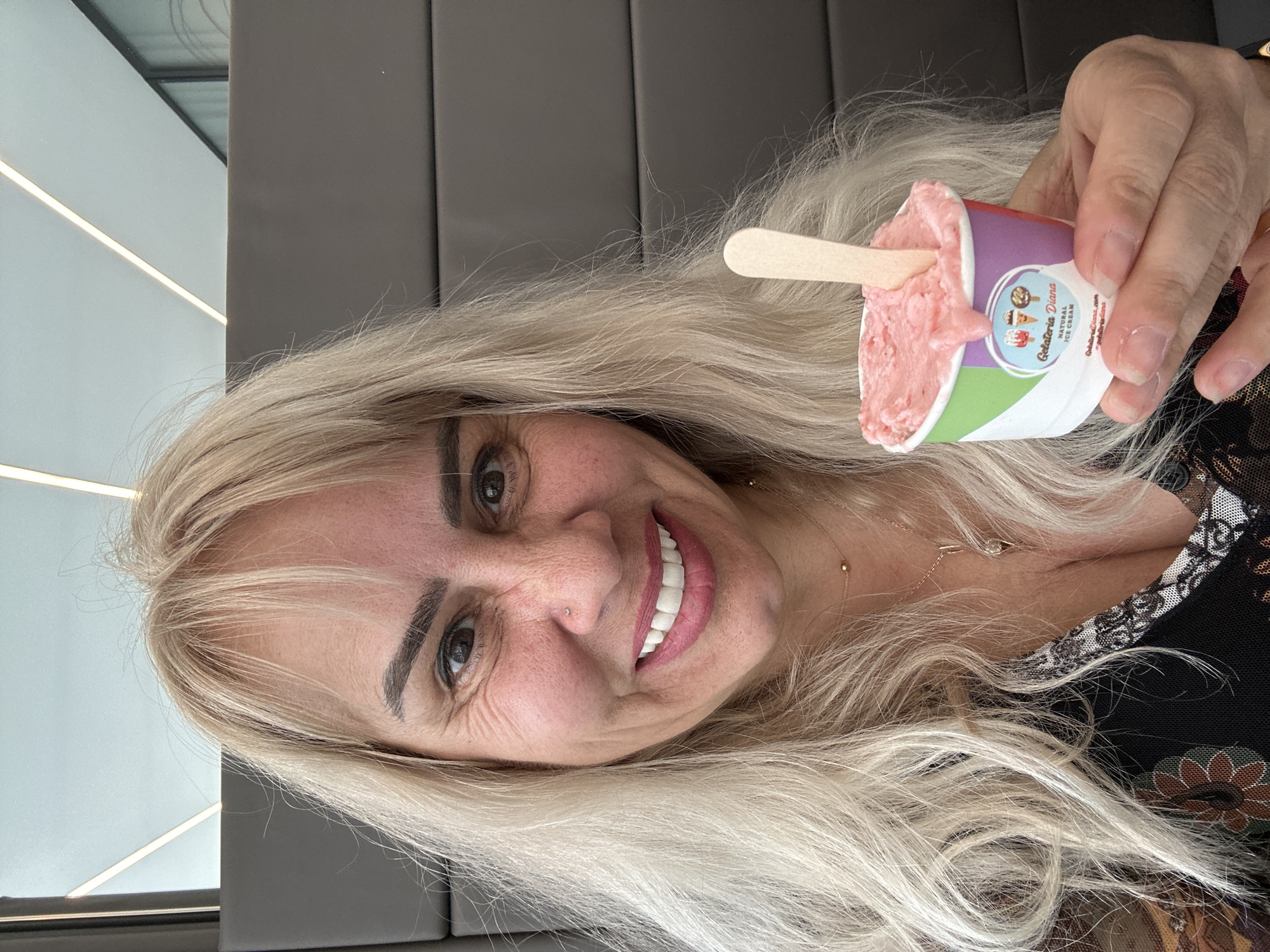 A close-up, brightly lit portrait captures a smiling woman holding a small cup of pink gelato. The woman has long, voluminous blonde hair, parted to the side, framing her face. She is smiling broadly, revealing bright white teeth, and has visible laugh lines around her dark eyes. Her eyebrows are dark and well-groomed, and she has a small nose piercing. She wears a dark top with intricate patterns and a lace trim at the neckline, and a delicate gold necklace with a small pendant is visible.
In her right hand, she holds a small, colorful paper cup filled with a light pink frozen dessert, presumably gelato or ice cream, with a small wooden spoon sticking out. The cup's label prominently displays "Gelateria Diana" with "NATURAL ICE CREAM" below it. Also visible are the website "GelateriaDiana.com" and the handle "@GelateriaDiana".
The background suggests a modern indoor setting, likely a cafe or restaurant. It features plain gray paneling behind her and a lighter-colored wall with horizontal light strips visible in the upper left corner. The image is taken from a slightly tilted perspective, giving a dynamic feel. The overall mood is one of happiness and enjoyment. This scene is set in Sofia, Bulgaria.