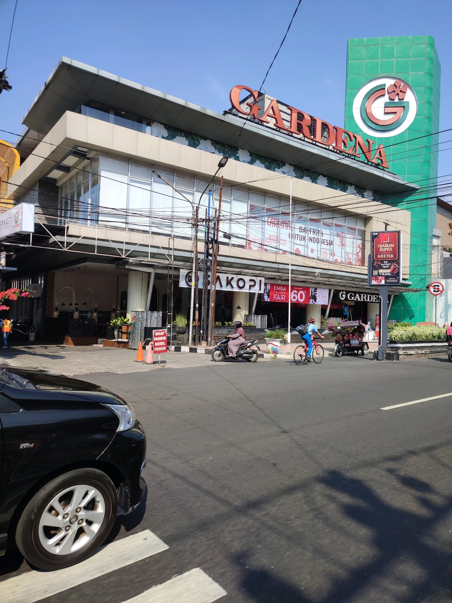 The image depicts a street scene in Magelang, Indonesia, in front of the GARDENA department store. The building, a multi-story structure with a large green tower featuring the GARDENA logo, dominates the background. The store's facade displays various signs, including "GARDENA" in large red lettering and advertisements for "CARDINAL," "EMBA FASHION FAIR 60," and "DJARUM SUPER KRETEK."
In the foreground, a black car is partially visible on the left, driving on a road with a pedestrian crossing. Several people are present: a person on a scooter to the left, another on a bicycle in the middle of the road, and a person on a motorized tricycle further to the right. Two orange traffic cones are placed on the sidewalk, accompanied by a sign that reads "MAAF PARKIR MOBIL PENUH" (Sorry, parking is full). The environment appears to be a sunny day, with clear blue skies.