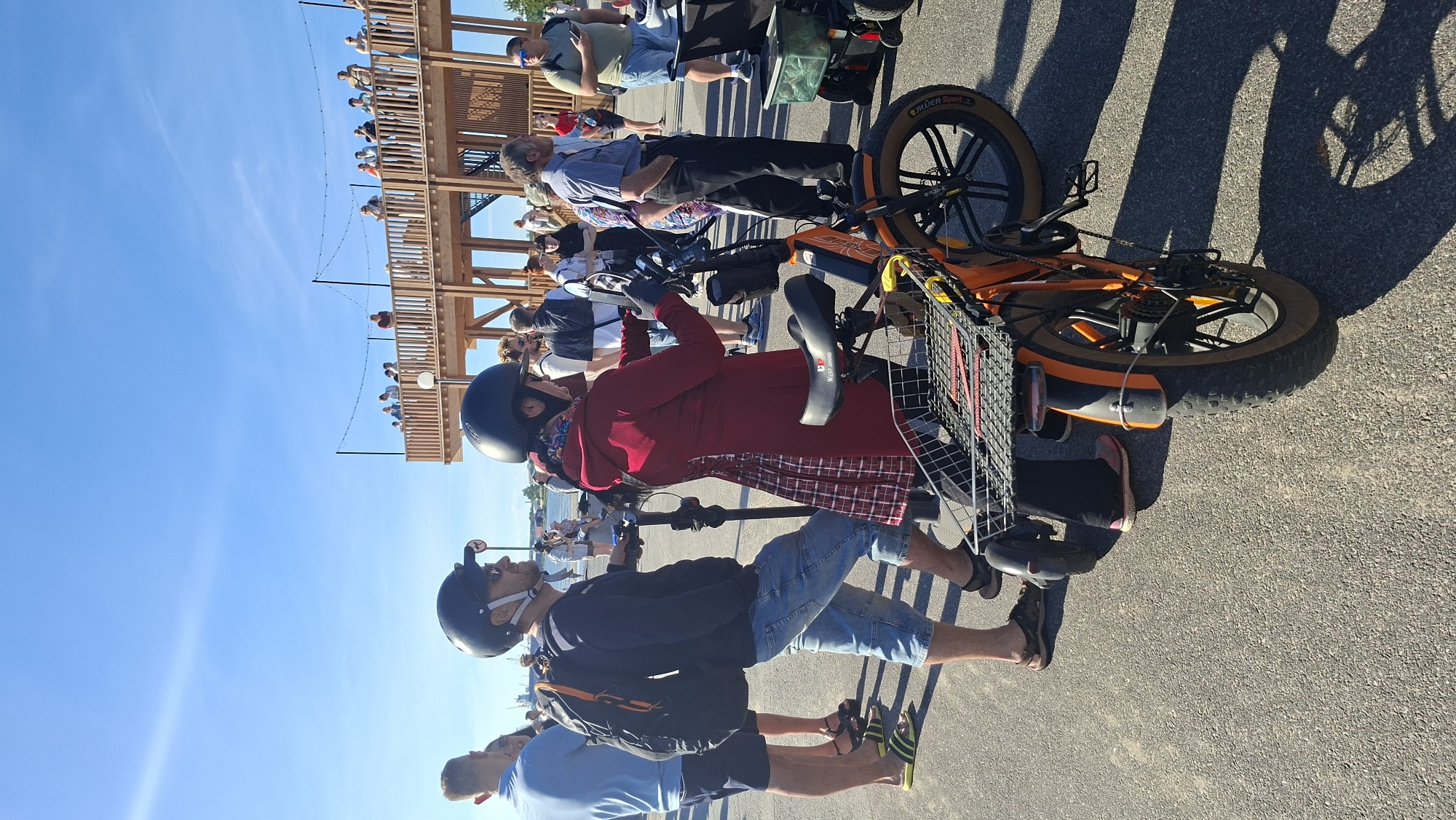 The image shows an outdoor scene with a group of people near a wooden structure, likely a pier or boardwalk, under a clear blue sky. In the foreground, two people are positioned next to electric scooters or bicycles. The person on the left, wearing a dark long-sleeved shirt, light blue denim shorts, and a black helmet, has a black backpack on. The person on the right is wearing a red coat and a black helmet, and is holding onto the handlebars of an orange electric bicycle with a basket in the front and rear. Another person wearing a blue polo shirt and dark shorts is visible to the left of these two. In the background, several people are standing on the wooden structure, which is decorated with string lights. The environment appears to be a public recreational area. Based on the architecture of the wooden structure and the general atmosphere, this scene is likely located in Tallinn, Estonia, possibly near the waterfront or a leisure district.