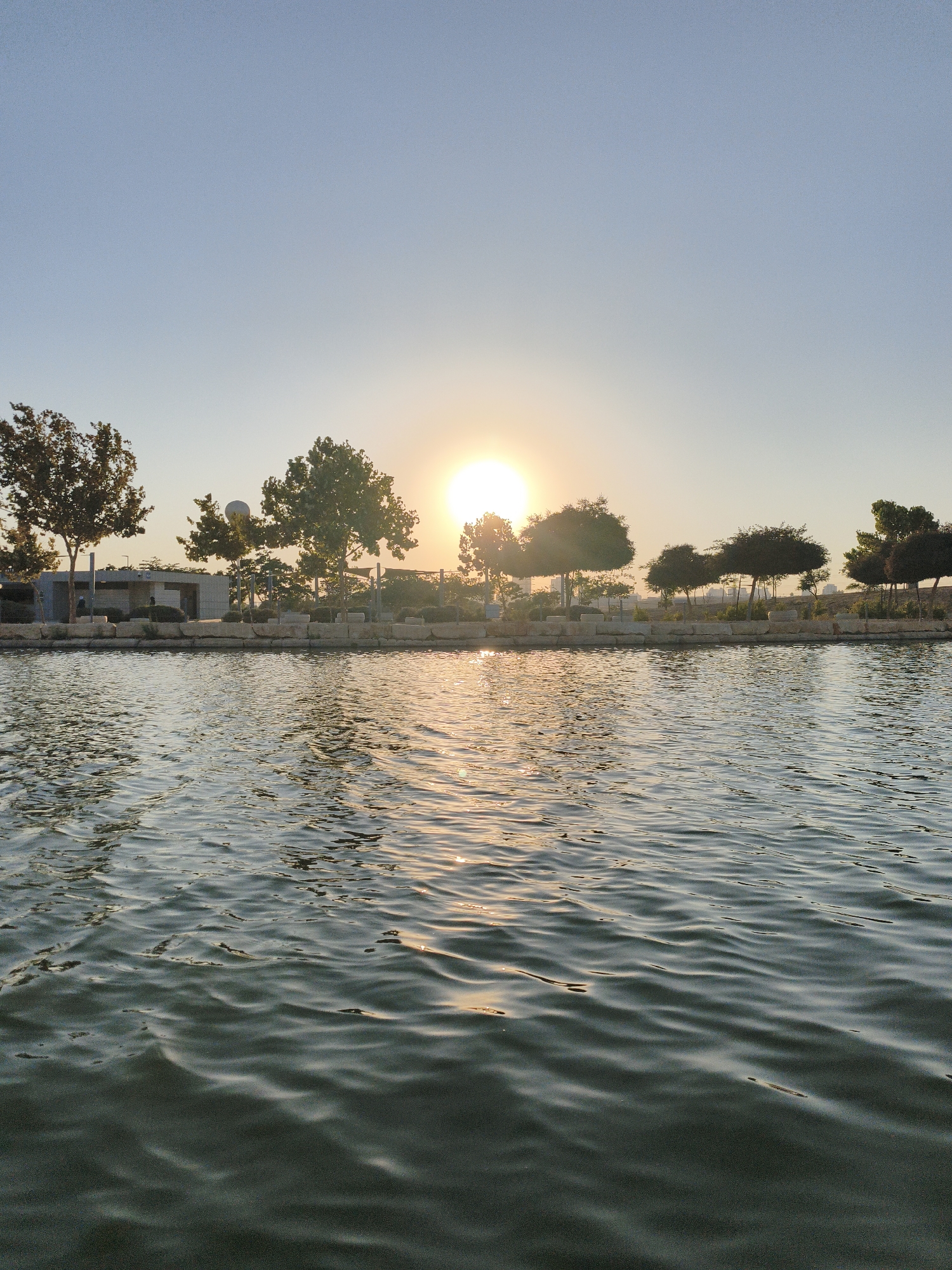 The image captures a serene waterscape at the Tel Aviv University campus in Be'er Sheva, Israel, likely during the late afternoon or early evening. A calm body of water dominates the foreground, its surface rippled by a gentle breeze, reflecting the golden hues of the setting sun and the clear blue sky above. Along the embankment, a line of trees and low-lying bushes are silhouetted against the bright sky. Behind the greenery, modern architecture, possibly part of the university buildings, is visible, along with a prominent spherical structure. The sun, low in the sky, casts a warm glow across the scene, its rays glinting off the water and illuminating the foliage. No people are discernible in the image.