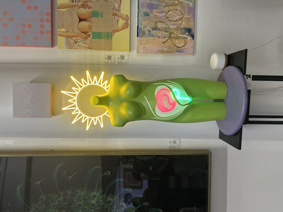A vibrant art display features a green, stylized torso mounted horizontally on a purple base. A neon sun radiates from the head area, while a glowing pink and green heart-shaped form is embedded in the abdomen. Above, an array of paintings, including one with pastel geometric shapes and another depicting a figure, adorn the wall. Below, a framed artwork with peacock imagery completes the composition.