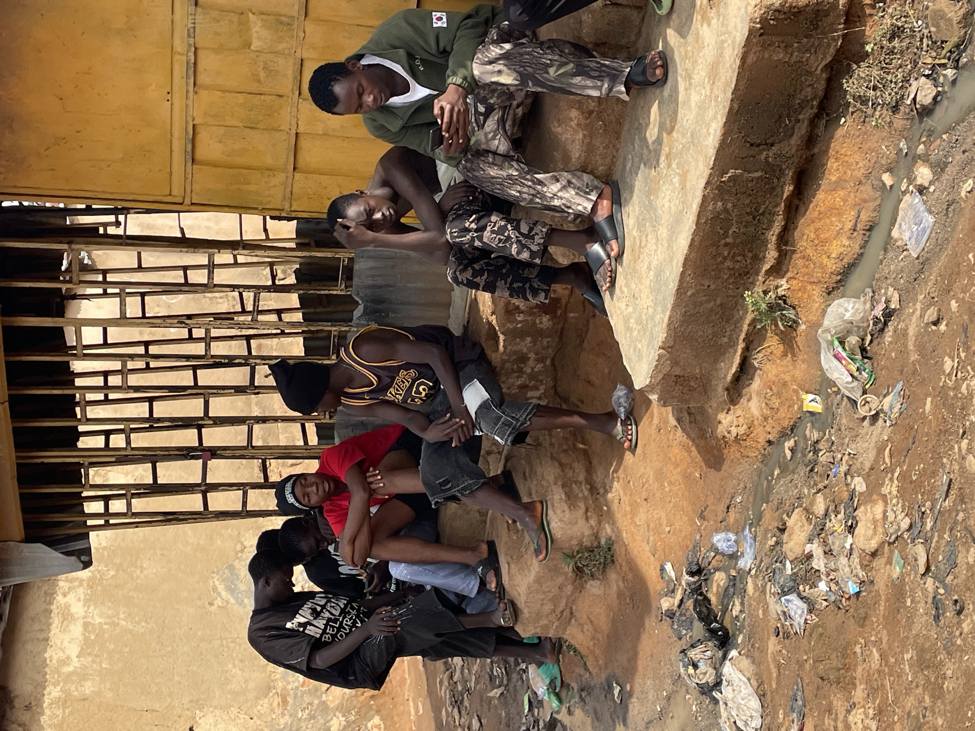 In Jos, Nigeria, several young men are sitting and standing near a concrete wall and building with a caged window, next to a dirt and gravel ground strewn with trash, rocks, and puddles. One man sits on a concrete step wearing an olive green jacket with a Korean flag patch, camouflage pants, and sandals, looking at his mobile phone. Another man next to him wears camouflage pants and sandals. Underneath them, a man in a Lakers jersey sits. Below him, two men wearing red and black are seated and interacting with each other. At the bottom of the image, a standing man wears a dark t-shirt and pants.