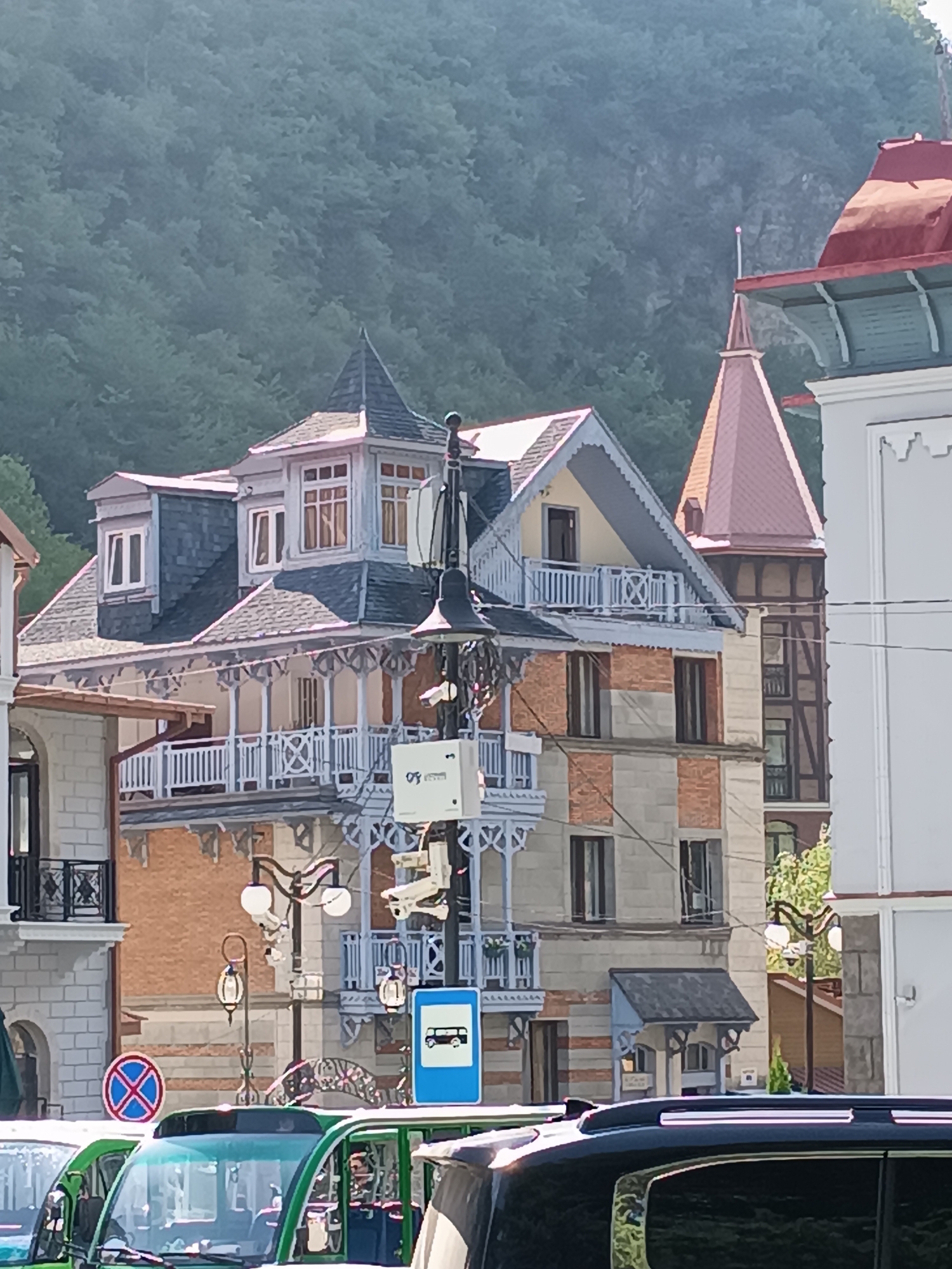 This image, captured in the city of Borjomi, Georgia, depicts a street scene with multiple buildings and vehicles. The architecture of the buildings appears to be in a European style, with ornate balconies, pitched roofs, and detailed facades. One prominent building features a multi-tiered structure with balconies adorned with white latticework, and a pointed turret with a red roof. Another building to its right has a prominent red roof and a tall, narrow tower with a pointed roof. The foreground is dominated by the tops of several vehicles, including a green vehicle with a visible windshield and a black vehicle. Several street lamps are visible, some with multiple globes. There are also traffic signs, including a blue sign with a bus icon and a red circular sign with a blue cross. The background shows a dense, green hillside covered in trees, indicating a natural landscape surrounding the urban area. The sky is bright, suggesting it is daytime.
