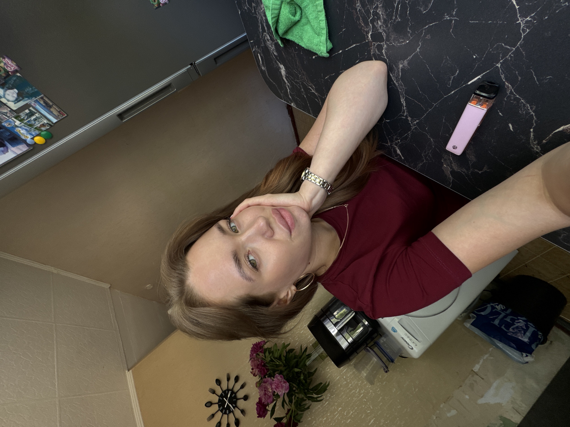 In the city of Chaikovsky, Russia, a young woman with long, light brown hair and green eyes is captured in a selfie. She is wearing a maroon t-shirt, a gold necklace, and a silver wristwatch. She has a gold hoop earring in her left ear. The woman is leaning her right hand on her face, her left arm resting on a black marble-patterned table. To her right on the table is a green towel and a pink electronic cigarette. In the background, a refrigerator with photos attached to it is visible, as well as a washing machine, a vase with pink flowers, and a decorative clock on the wall. The environment appears to be a domestic setting, likely a kitchen.