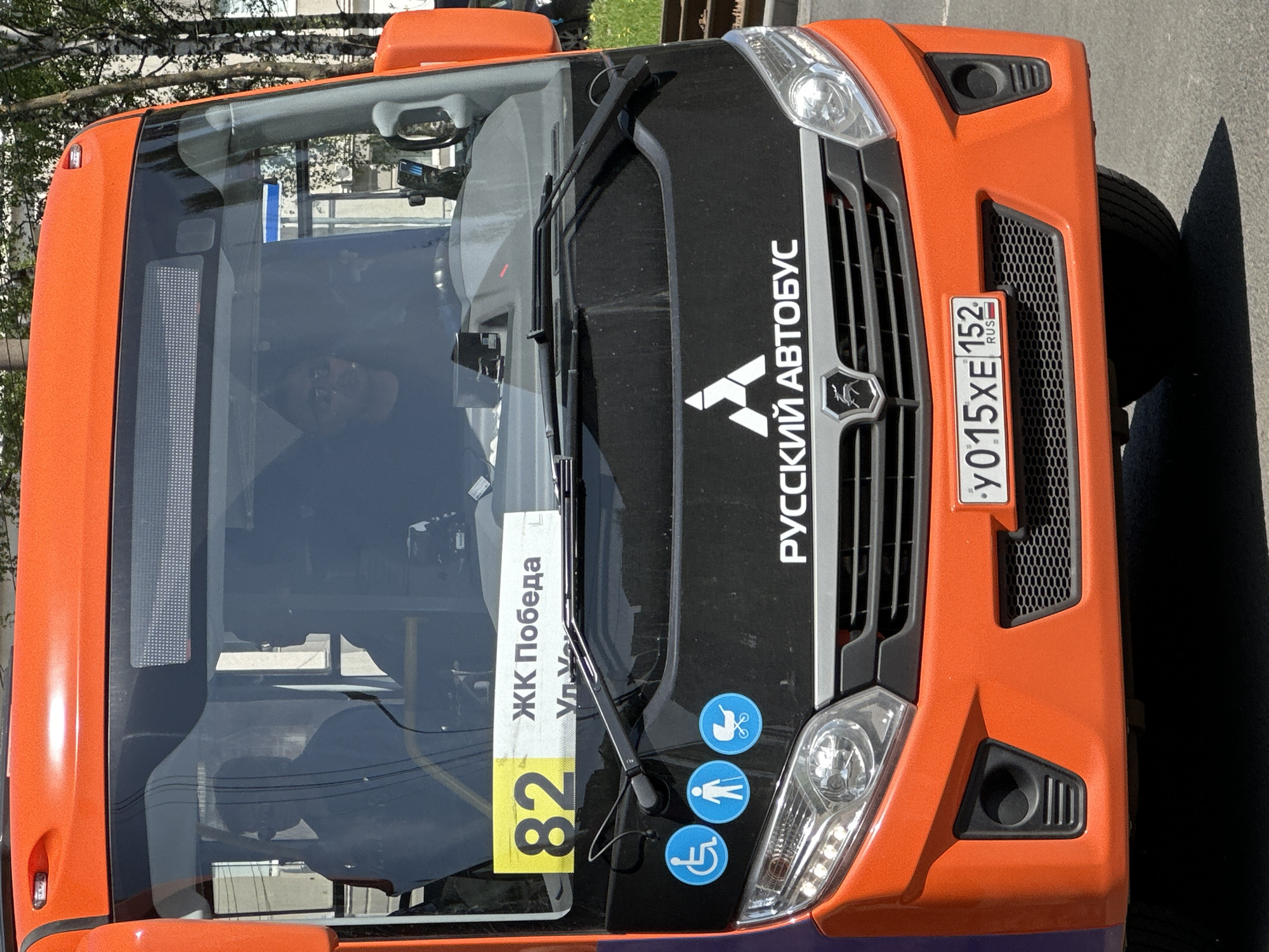 The image shows the front of an orange "Russian Bus" (Русский автобус) bus, route number 82. The route identifier is "ЖК Победа" (Residential Complex Pobeda). The bus displays a license plate: "y015XE 152 RUS". It also shows the international disability and pedestrian symbols. The location appears to be Nizhny Novgorod, Russia, judging by the bus name and the license plate region code (152).