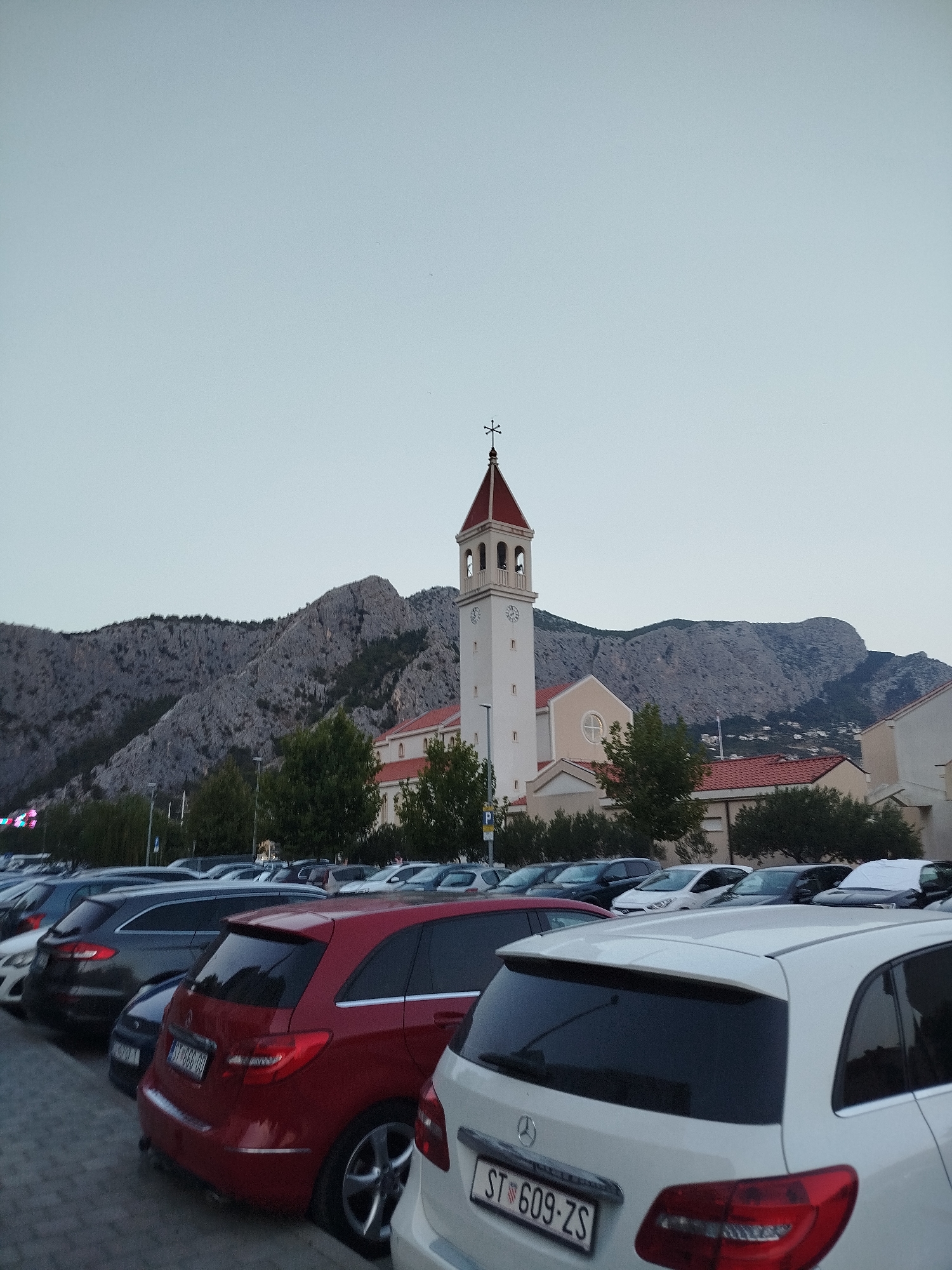 A parking lot is filled with cars in front of a building with a bell tower, likely a church, and a rocky mountain range in the background. The sky is overcast and pale. The church has a tall, white rectangular tower with a red-tipped spire and bells visible in arched openings. Attached to the tower is a building with a red-tiled roof and light-colored walls. The mountain range appears rugged and barren, with some sparse vegetation on its lower slopes. Several cars are parked in rows, including a red hatchback and a white hatchback in the foreground. A license plate on the red car reads "ST 666-GG". The scene is likely in Omiš, Croatia, given the presence of the church and mountains, common features of coastal Croatian towns.