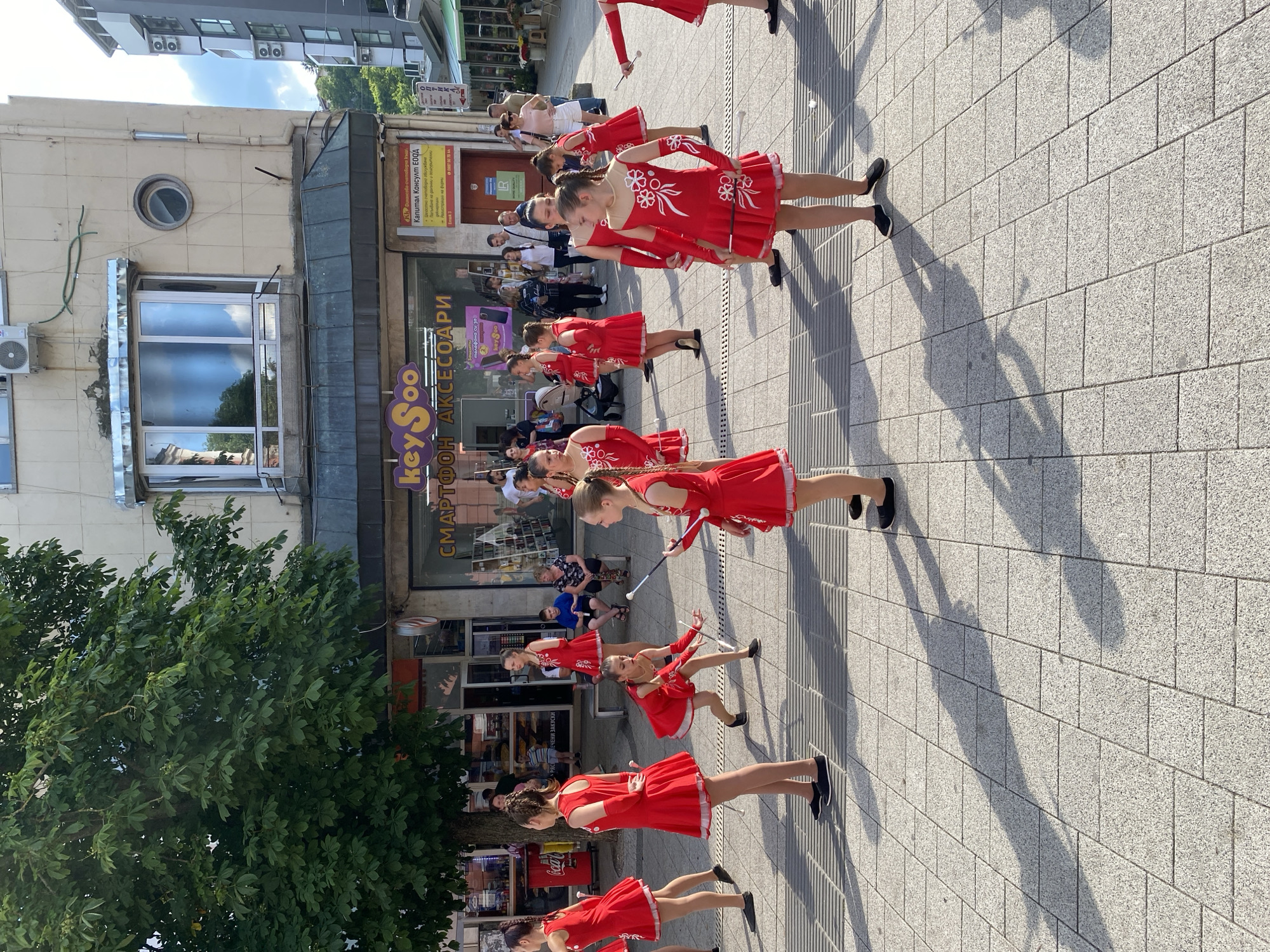 The image captures an outdoor daytime scene in Pleven, Bulgaria, on a brightly lit pedestrian square. Approximately ten young girls, appearing to be pre-teens, are dressed in identical short-sleeved red dresses with white trim and decorative patterns, and dark slip-on shoes. Most hold silver and black batons, suggesting a majorette or twirling performance. One girl in the center-left foreground is captured mid-squat with her baton extended forward, while others stand in various poses.
To the left, a large, leafy green tree casts a shadow across part of the paved area. In the background, a light-colored multi-story building features several ground-floor shops. Visible signage includes a prominent purple and yellow "keySoo" logo with the Bulgarian text "СМАРТФОН АКСЕСОАРИ" (Smartphone Accessories) beneath it. To the right of keySoo, a yellow sign displays "Капитал Консулт ЕООД" (Capital Consult Ltd.) in red Cyrillic text. The upper floors of the building feature a bay window, a small circular window, and air conditioning units. Further to the right, modern grey buildings with multiple external AC units are visible against a partly cloudy blue sky.
Several adult bystanders observe the scene; some are seated on a bench to the left, while others stand further back. The ground is paved with light grey rectangular interlocking stones, interspersed with metallic grates. Long, distinct shadows of the performers and onlookers stretch across the pavement, indicating strong sunlight from the right.