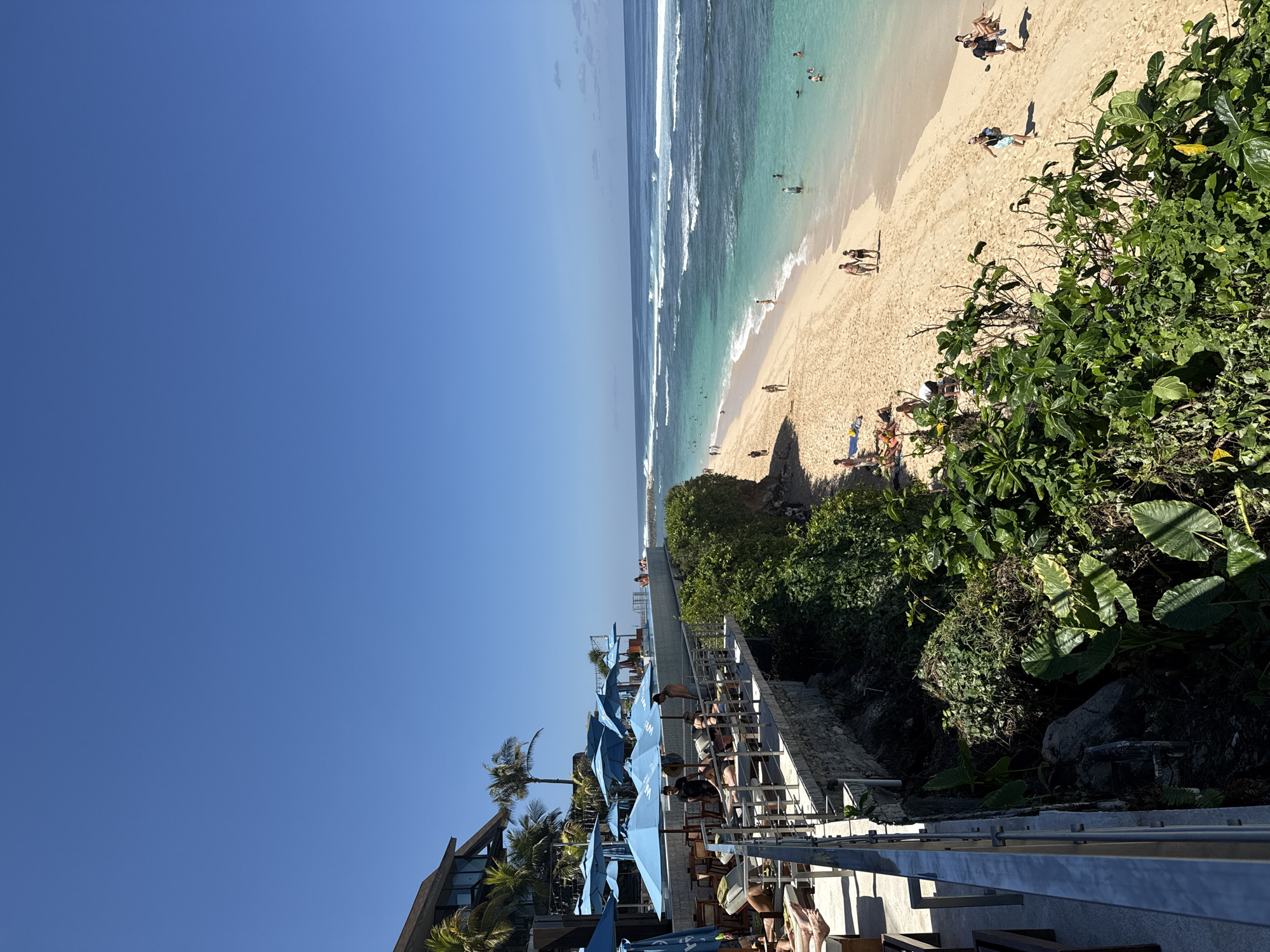 An aerial view shows a tropical beach in Kuta Selatan, Indonesia, on a clear, sunny day. The foreground features a resort with a restaurant area set up with blue umbrellas and lounge chairs, populated by a few patrons. A metal railing separates the resort area from a steep, vegetation-covered cliff. The beach itself is a strip of golden sand bordering a turquoise ocean with moderate waves. Several people are visible on the beach, some walking and others swimming or wading in the water. The overall scene depicts a vibrant and relaxed atmosphere at a popular tourist destination.