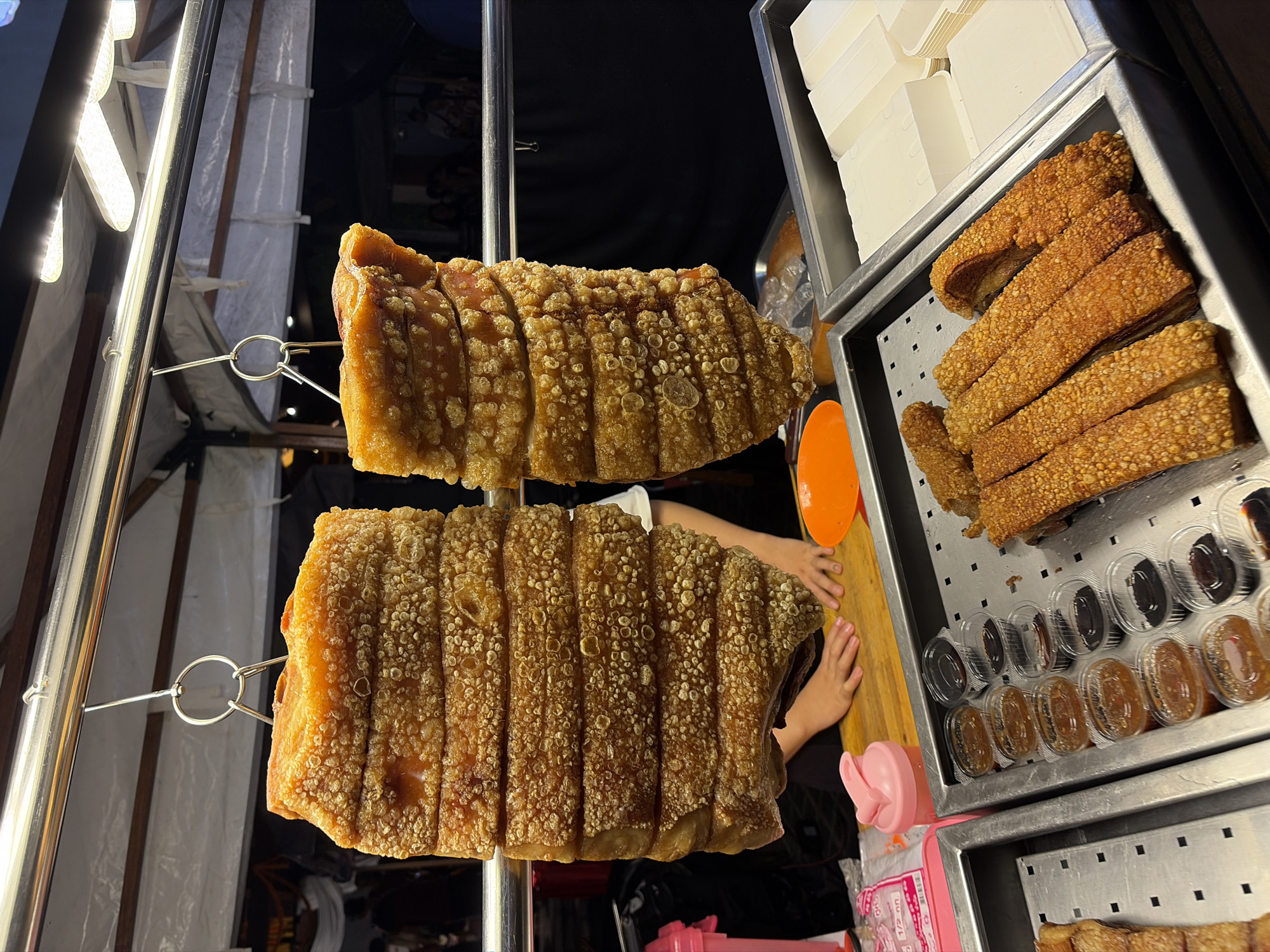 In the city of Chiang Mai City Municipality, Thailand, at a food stall, two large blocks of crispy pork belly are displayed hanging from a metal rack. The pork belly is cut into thick, rectangular slices, revealing layers of meat and fat, and is covered in a golden-brown, bubbly, and crispy skin. Below the hanging pork, a metal tray holds several rows of small plastic containers filled with dipping sauces, appearing to be of varying shades of brown and amber. Adjacent to this tray is another metal container filled with rectangular portions of what appears to be pork crackling or more crispy pork. To the right, white takeaway containers are stacked, ready for use. A person's hands are visible on a wooden cutting board, suggesting preparation or serving activities. The background is dark, with hints of a white tent-like structure and indistinct figures, indicating an outdoor market or street food setting. The lighting suggests it is evening or night.