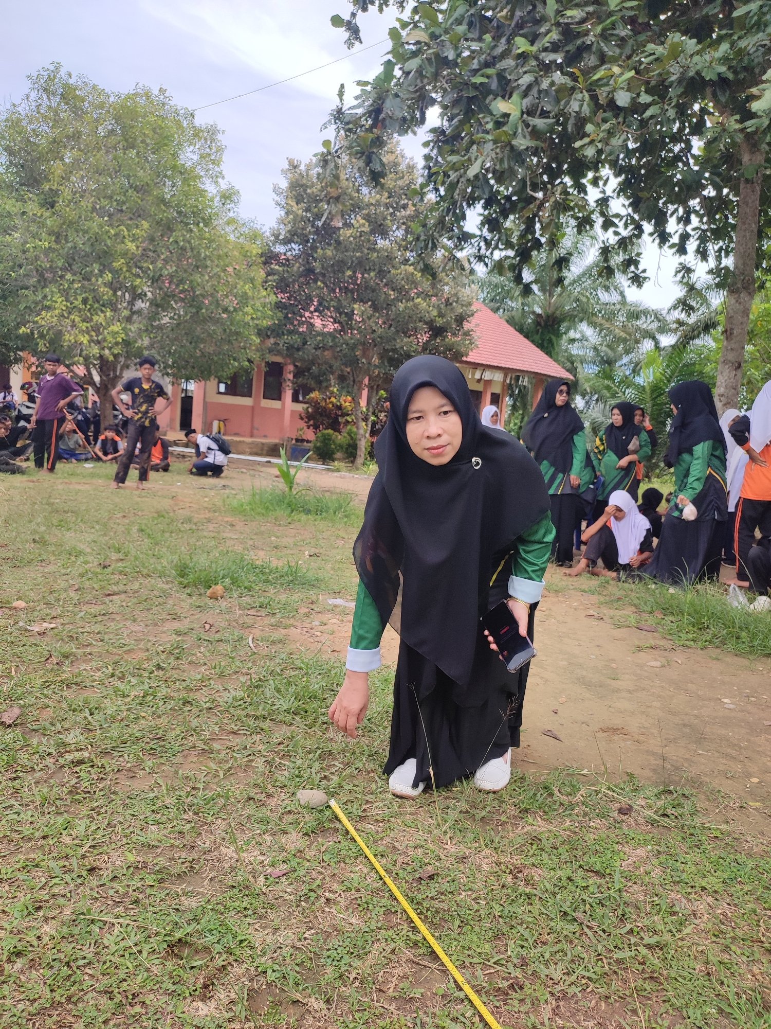 A woman in a black hijab and a green and black outfit is positioned in the foreground of this outdoor scene. She is holding a smartphone in her right hand and appears to be bending down to interact with a measuring tape and a small stone on the ground. Behind her, a group of people, some in similar uniforms, are gathered. They are on a dirt path bordered by grass and trees. In the background, a building with a red roof is visible. The sky is overcast. The scene appears to be taking place in a schoolyard or similar communal area, possibly during an outdoor activity or event. The location is specified as Blangriek, Indonesia.