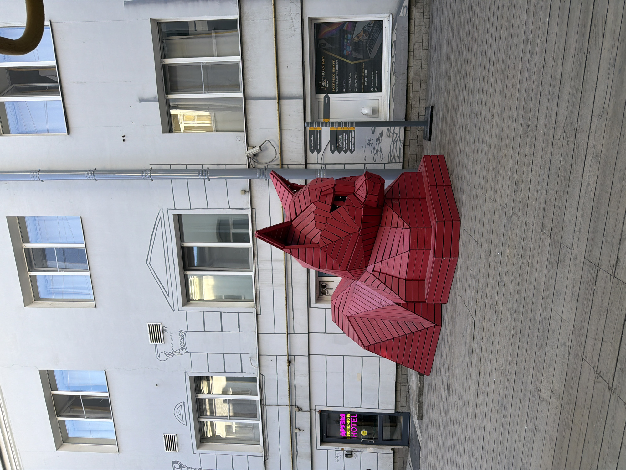 This is a full-shot, eye-level, outdoor photograph of a red, angular, geometric sculpture of a cat in an urban courtyard. The sculpture is positioned in the foreground, angled slightly to the left, and is made of numerous red wooden planks. To the left of the cat sculpture is a glass door with pink neon lettering that reads "ДРУЗЬЯ ЭТО ТО, ЧТО ТЕБЯ HOTEL" and a yellow hotel sign. Behind the sculpture is a light-colored building with multiple windows. To the right of the cat sculpture is a directional signpost with yellow signs pointing in various directions, and a window displaying advertisements. The courtyard floor is made of weathered grey wooden planks. The scene appears to be in Saint Petersburg, Russia, as indicated by the neon signage on the door. There are no people visible in the image.