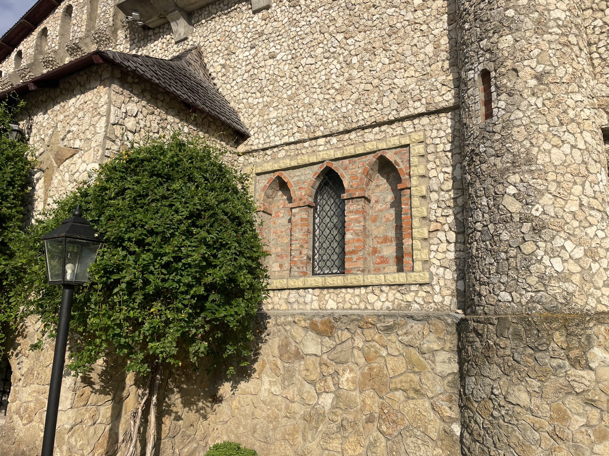 The image shows a section of a stone building exterior, possibly a castle or historical structure, in Colibași, Moldova. The wall is constructed from rough-hewn stones of varying sizes and colors, primarily in shades of beige and light brown. There is a prominent arched window with red brick detailing surrounding it, and a black metal grate covers the window. To the left of the window, a lush green bush or tree is visible, and a black lamppost with a glass lantern stands in front of it. The building appears to have a dark-tiled roof section visible in the upper left corner. The lighting suggests it is daytime, with sunlight casting shadows.