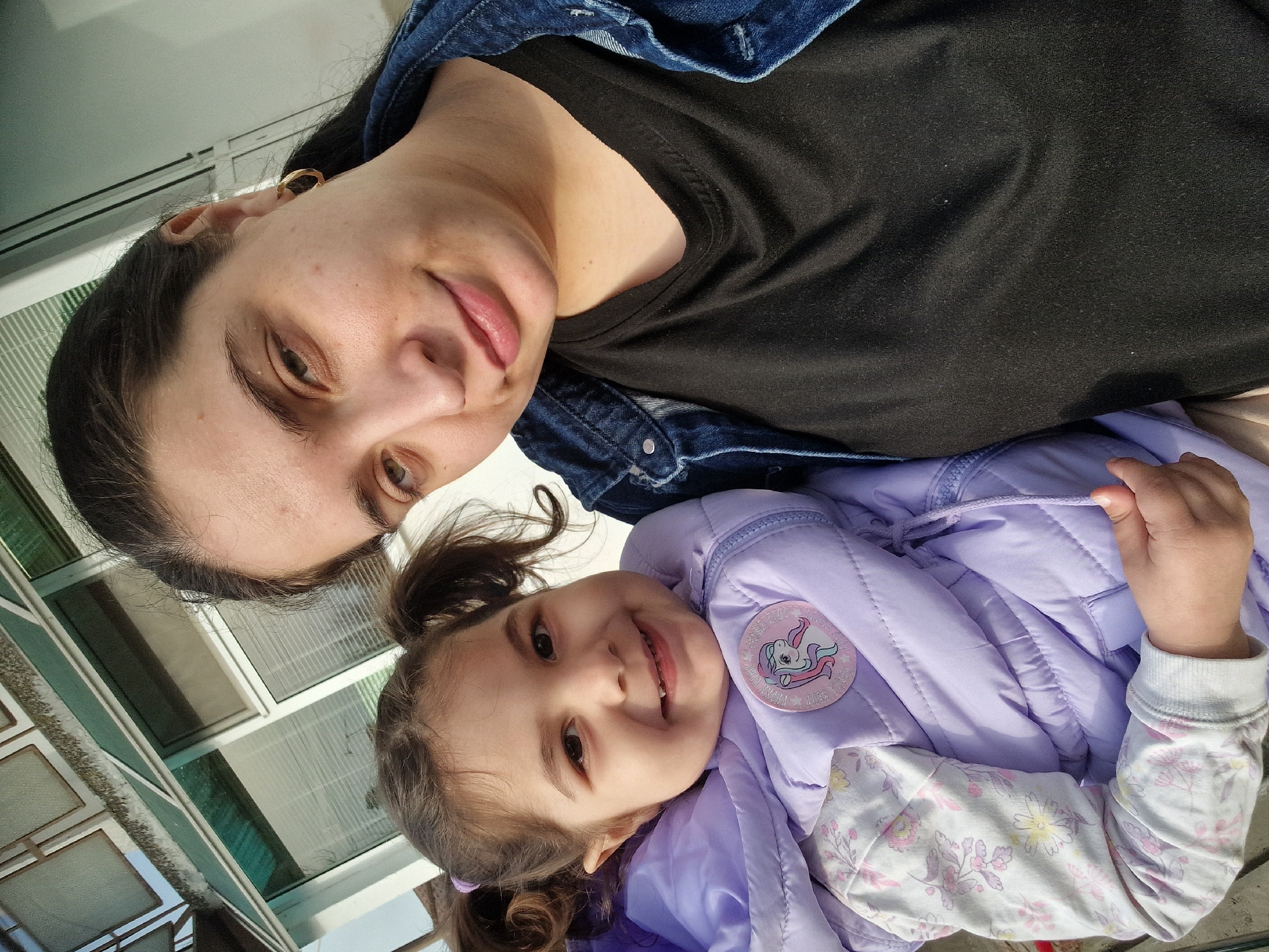 A medium shot depicts a woman and a young girl in front of a window, likely outdoors in an unknown city and state. The woman has dark hair pulled back, and wears an earring, a black shirt, and a denim jacket. She looks towards the camera with a slight smile. The young girl, also with dark hair in pigtails, wears a light purple puffer jacket over a floral shirt. A sticker of a pink unicorn is visible on her jacket. Both the woman and girl seem to be looking at the camera. A window is behind them.