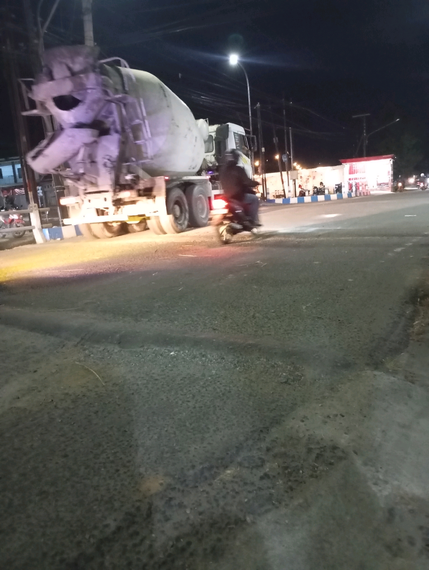 The image depicts a nighttime street scene in Pacitan, Indonesia, likely at an intersection. A large cement mixer truck is prominently featured on the left side of the frame, illuminated by its headlights and streetlights. Its drum is angled upwards, suggesting it is either en route to or from a construction site.
In the middle ground, a person is riding a motorcycle, moving away from the camera and towards the right. The motorcycle's taillight is visible, casting a red glow. Behind the motorcycle, several other motorcycles are parked or in motion near a brightly lit building complex. This complex appears to be a commercial establishment, possibly a roadside cafe or shop, with red and white signage.
The environment suggests an urban or semi-urban setting at night. Streetlights are visible, casting pools of light on the asphalt road, which appears to be wet in some areas, reflecting the light. Power lines are strung across the dark sky. The road surface shows signs of wear and tear, with some tire marks visible. The overall atmosphere is that of a typical busy intersection at night in an Indonesian city.