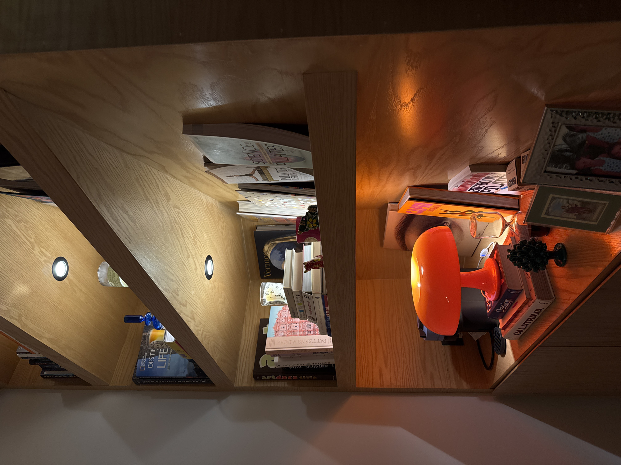 The image shows a close-up view of a bookshelf. The shelves are filled with a variety of items, including books, bottles, picture frames, and a bright orange lamp. The bookshelf is made of light-colored wood and is illuminated by built-in spotlights. The books have titles such as "Science," "artdeco style," "Design," "CALATRAVA," "DESTIN LIFE," "PATTERNS & INDIA" and "1000 PLACES TO SEE BEFORE YOU DIE." There is a bottle with a blue stopper in the lower-left section of the shelf. One shelf has small framed pictures. The image does not contain any information about Puebla, Mexico.