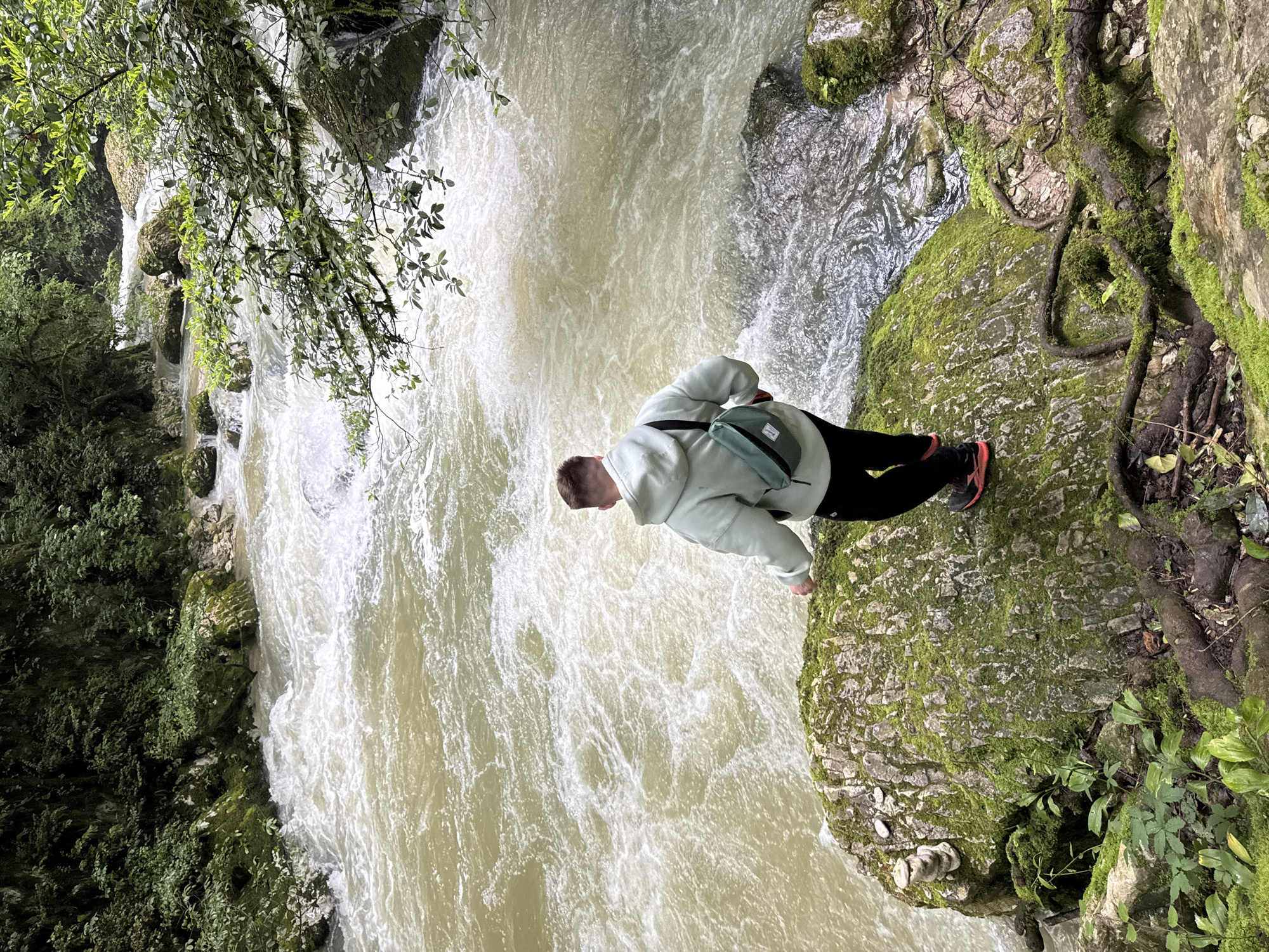 A person stands on a moss-covered rock outcropping, viewing a turbulent, milky-colored river flowing through a rocky gorge. The person wears a light-colored hooded sweatshirt, dark pants, and orange and black running shoes, with a small green bag slung across their body. Vegetation clings to the rocks on either side of the river, including a tree branch with leaves hanging into the scene from the upper left. The image is taken from above, creating a slightly distorted perspective. The location is Tsabal, Abkhazia.